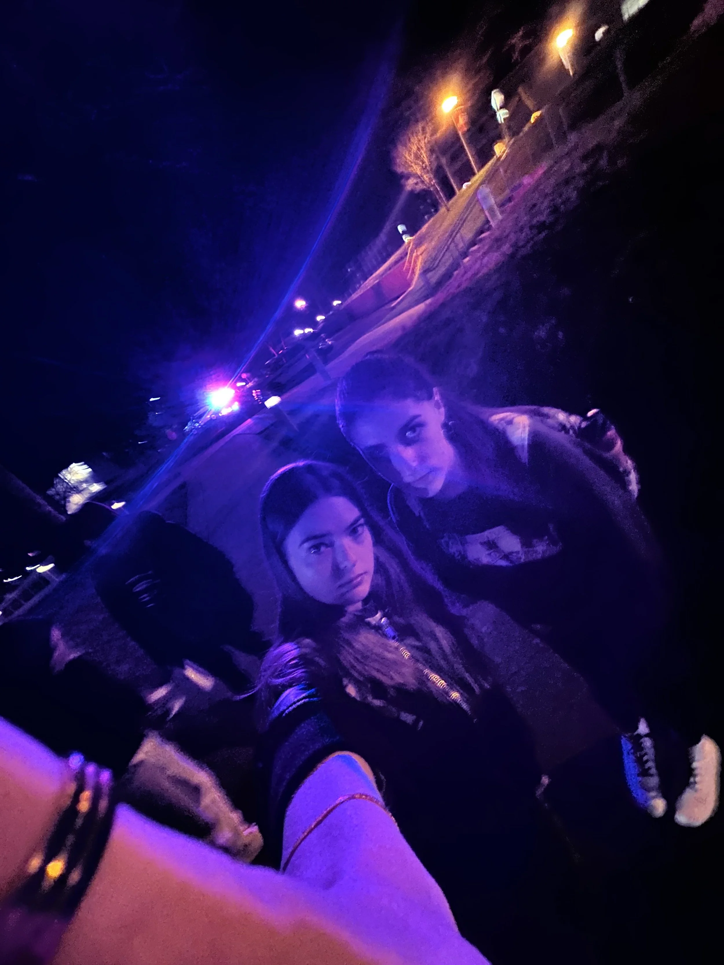 Two young women taking a selfie at night with blue and orange streetlights in the background. They are outdoors, near a sidewalk, with trees and cars visible in the distance.