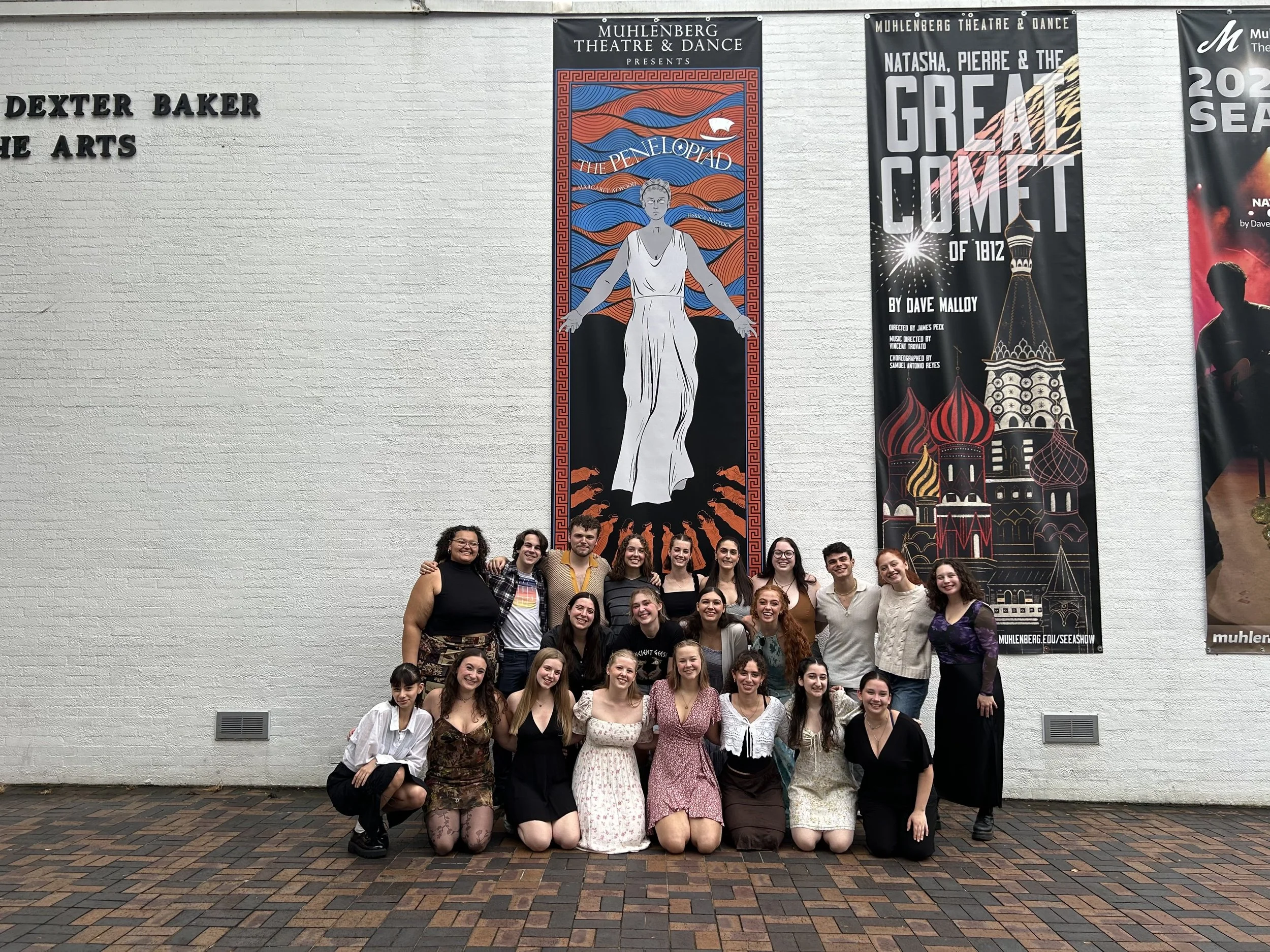 Group of people posing in front of posters at Muhlenberg Theatre & Dance, with a white brick wall and brick pavement, smiling and standing close together.