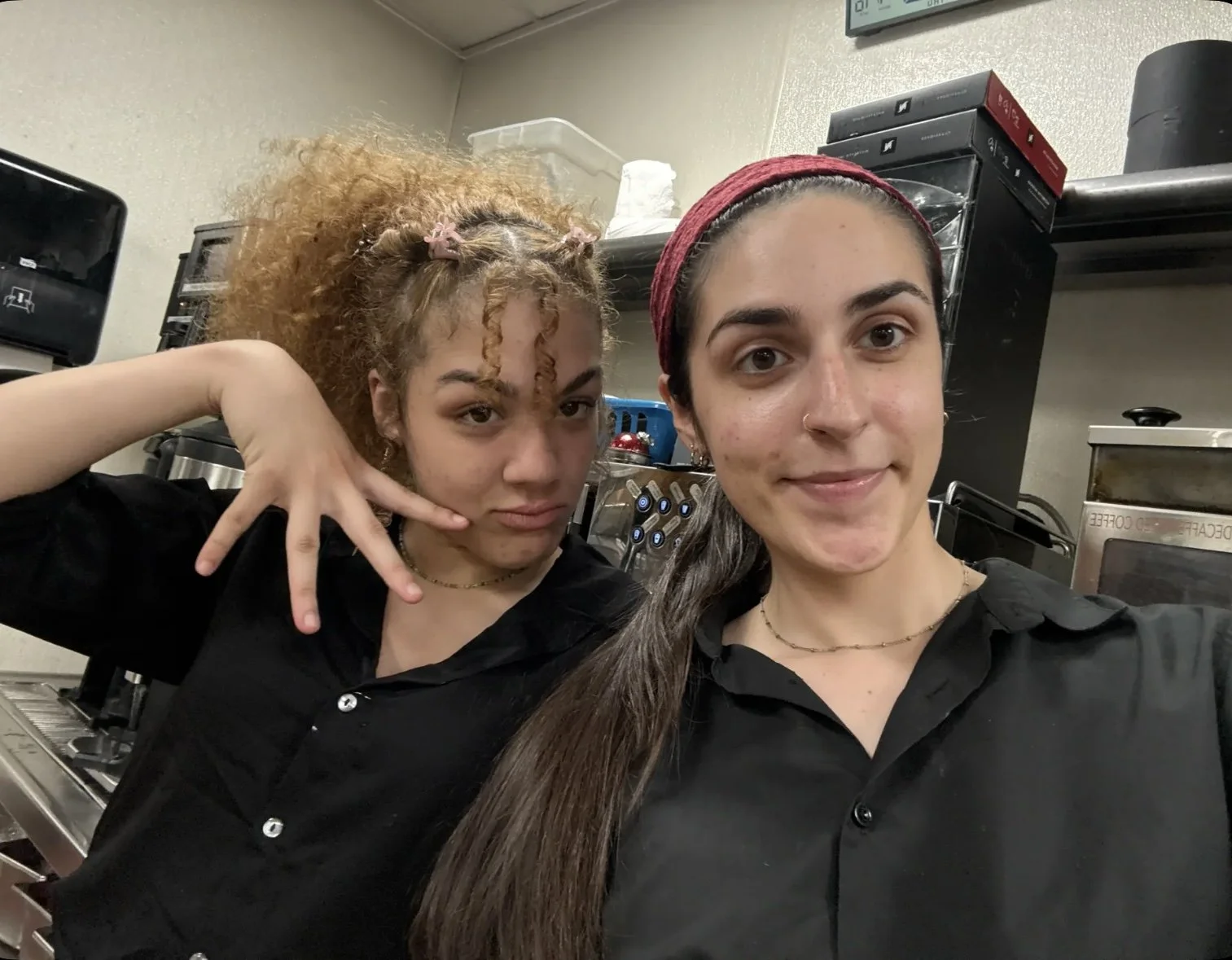 Two young women wearing black shirts taking a selfie in a kitchen or break room with coffee machines and storage containers behind them.