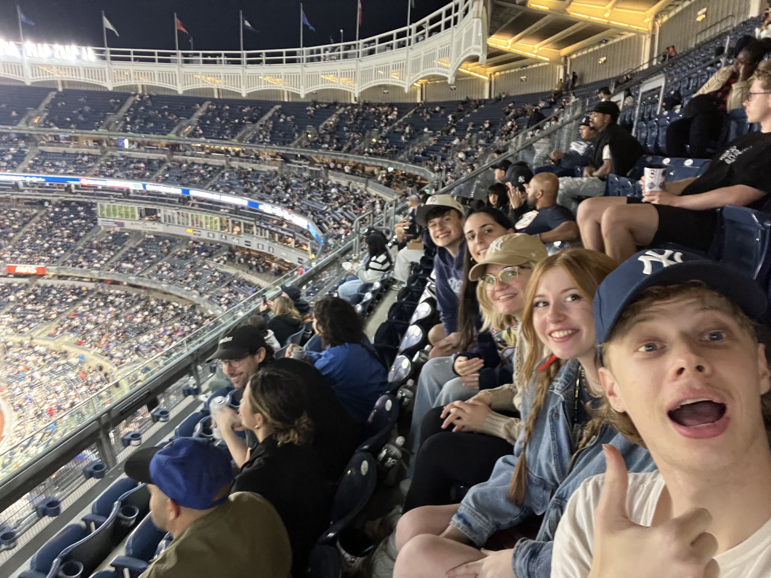 People seated in a stadium, watching an event, with some smiling and taking selfies, during the evening.