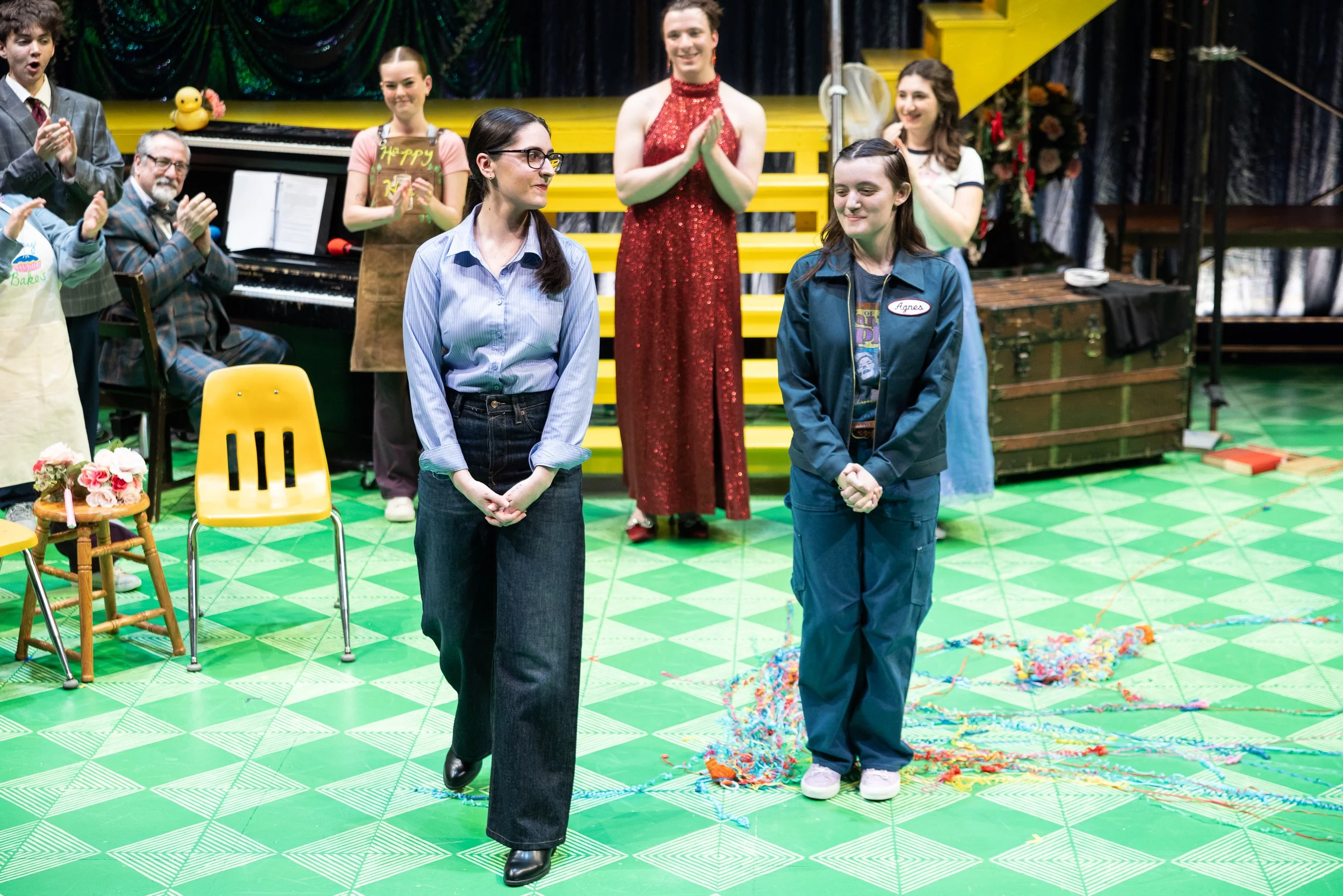 A group of people standing on a green patterned floor, with three people in the foreground. One woman is wearing glasses and a light blue button-down shirt, and another young woman is wearing a dark jacket. The background features a stage with a yell