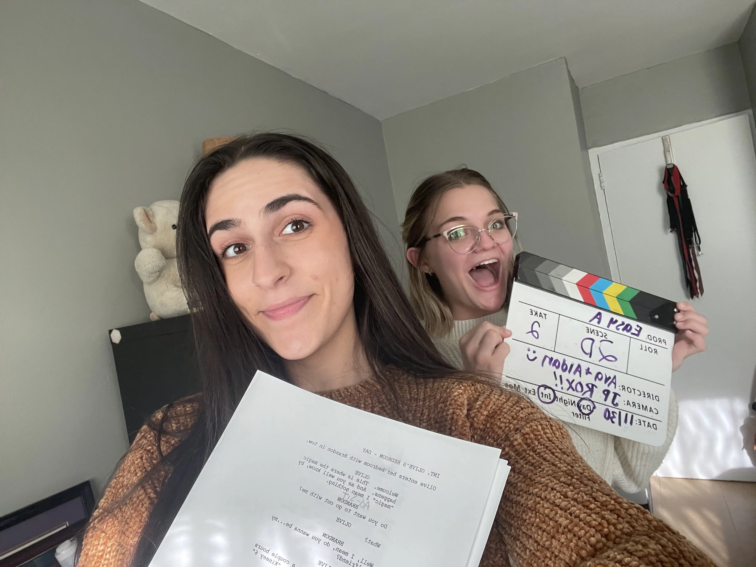 Two women smiling at the camera; one holding a clapperboard with various notes and the other holding a script. Behind them, a teddy bear is on a shelf, and part of a computer is visible in the foreground.
