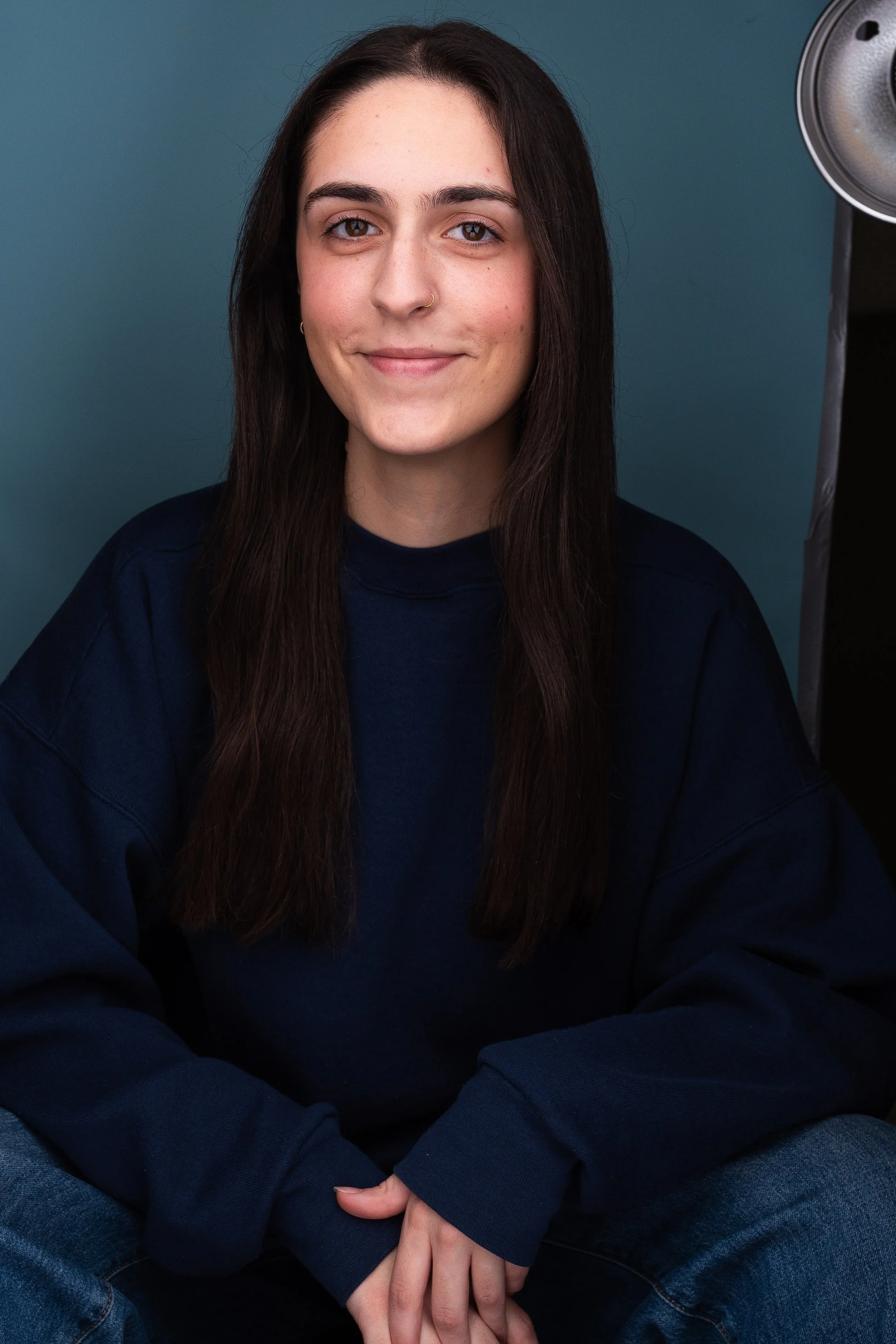 A young woman with long dark hair, wearing a navy blue sweatshirt, sitting with her hands folded on her lap, smiling gently at the camera, against a plain grey background.