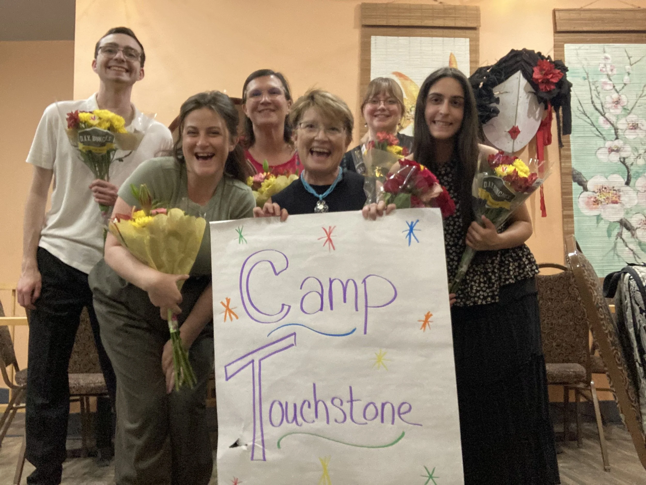 A group of seven smiling people holding flowers and a colorful handwritten sign that says "Camp Touchstone" in a decorated room with Asian-themed artwork and decor.