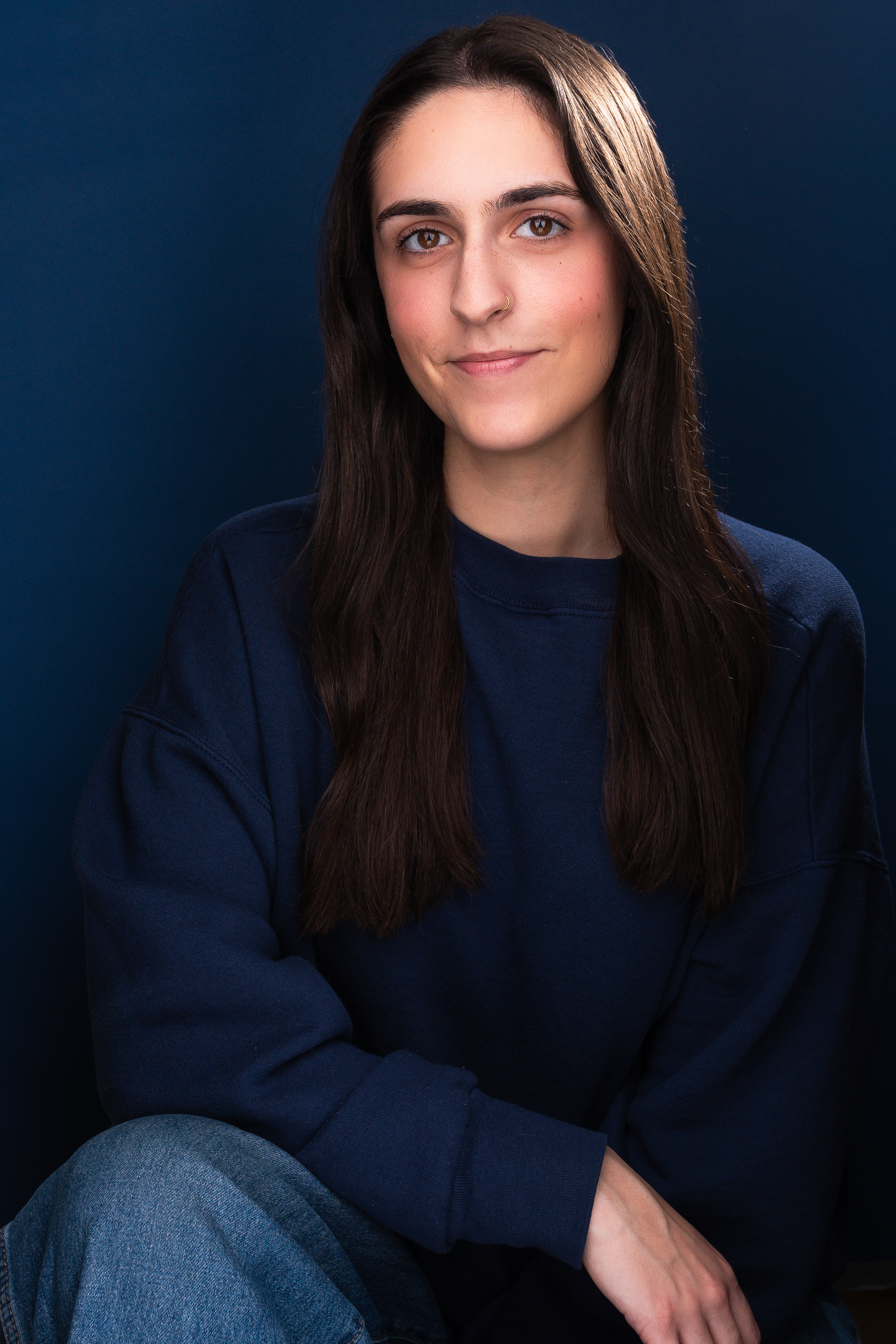 Portrait of a young woman with long dark hair, wearing a navy blue sweatshirt and blue jeans, sitting against a dark background.