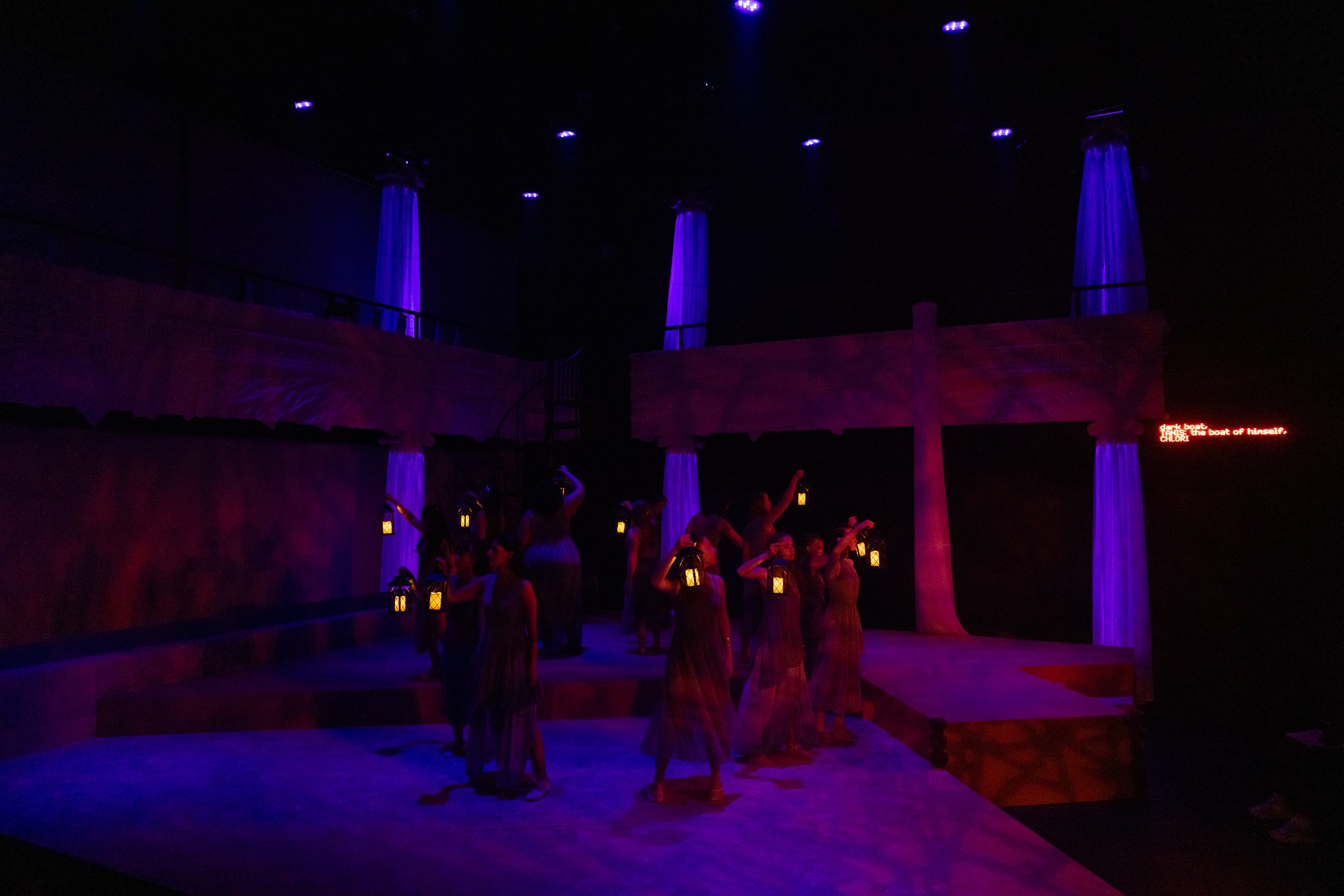 A dark indoor setting with purple lighting illuminating four tall pillars with flowing fabric. There are people, many holding small lanterns, standing around the pillars in dresses. A red digital sign displays text in the lower right corner.