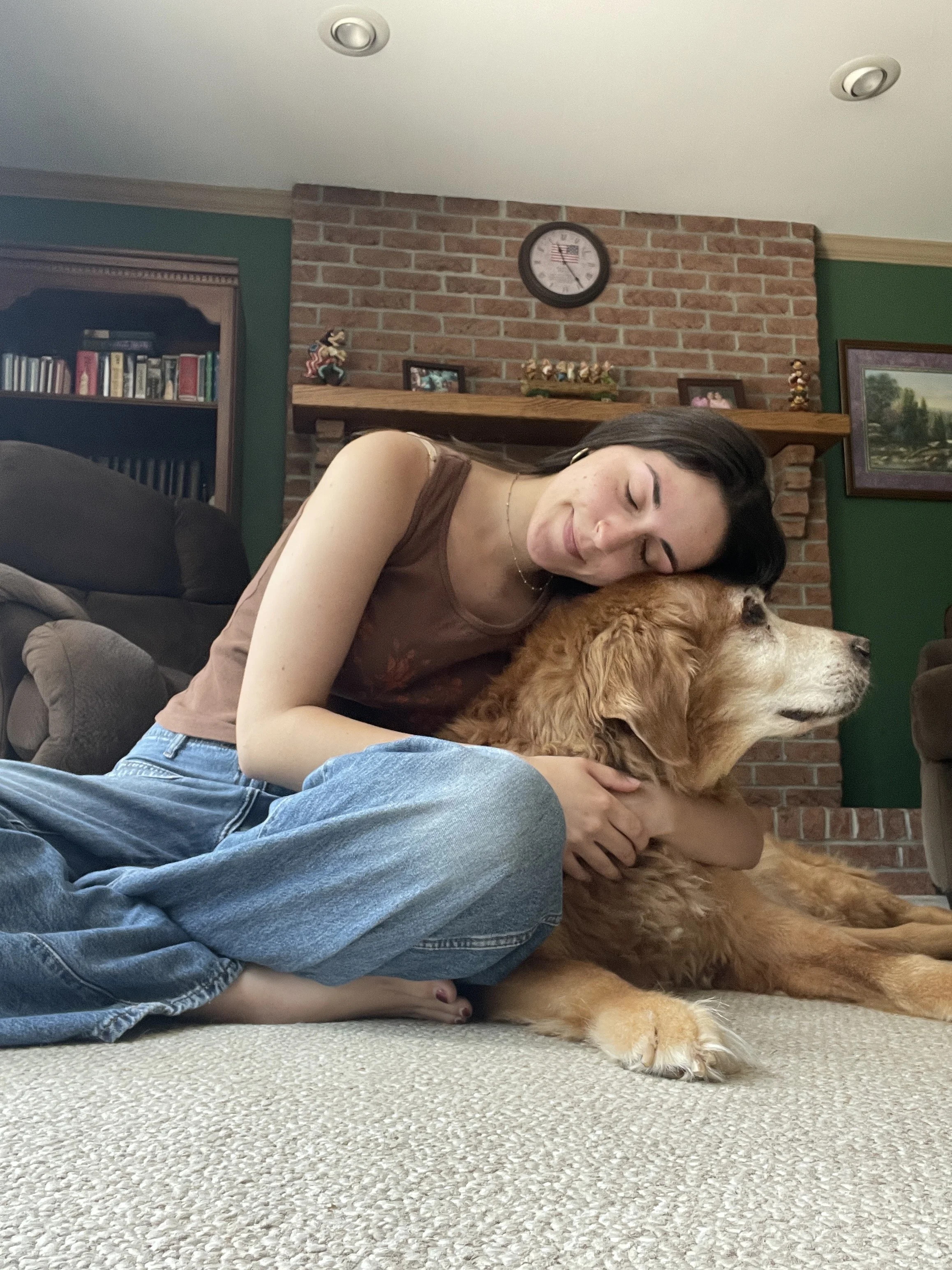 A woman with dark hair hugging a large golden retriever dog on a carpeted floor in a living room with a brick fireplace and a wooden mantel decorated with small figurines and framed photos.