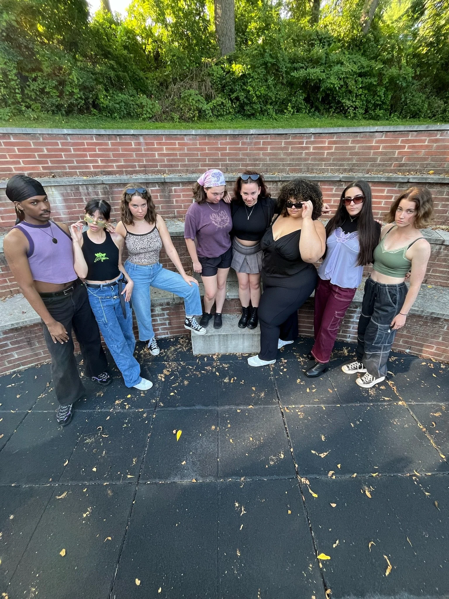 A group of nine women posing together outdoors on a sidewalk in front of a brick wall and lush green trees.