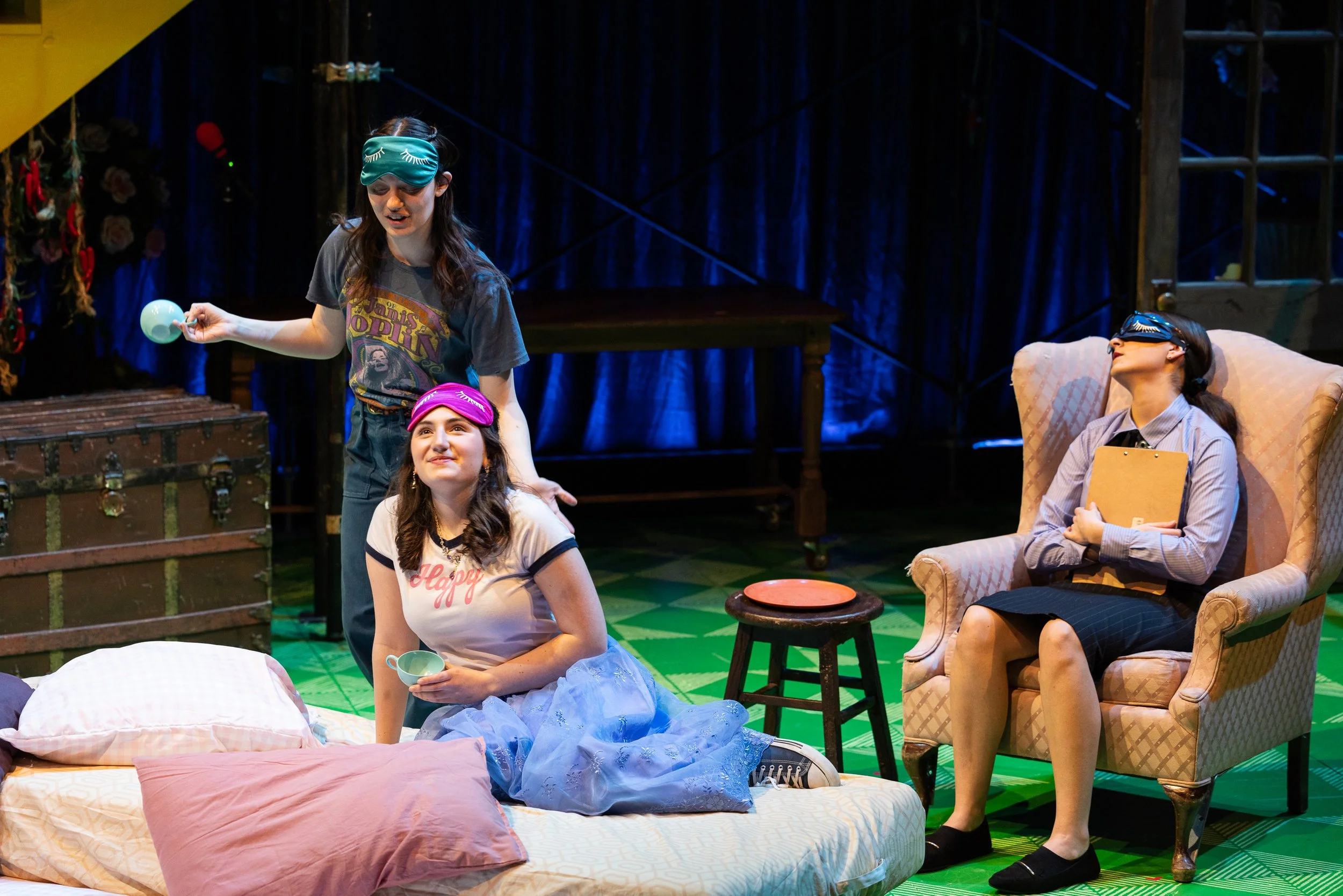 The scene depicts four women in a theatrical production, with two seated on the floor and one on an armchair, and one standing. The woman on the floor in front is holding a teacup, wearing a white t-shirt with 