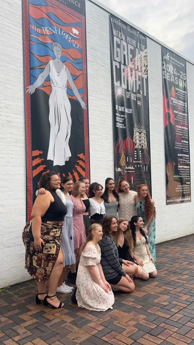 Group of people posing for a photo in front of large theater banners, including one with an illustration of a woman in a white dress and flowing hair, and others advertising performances and events.