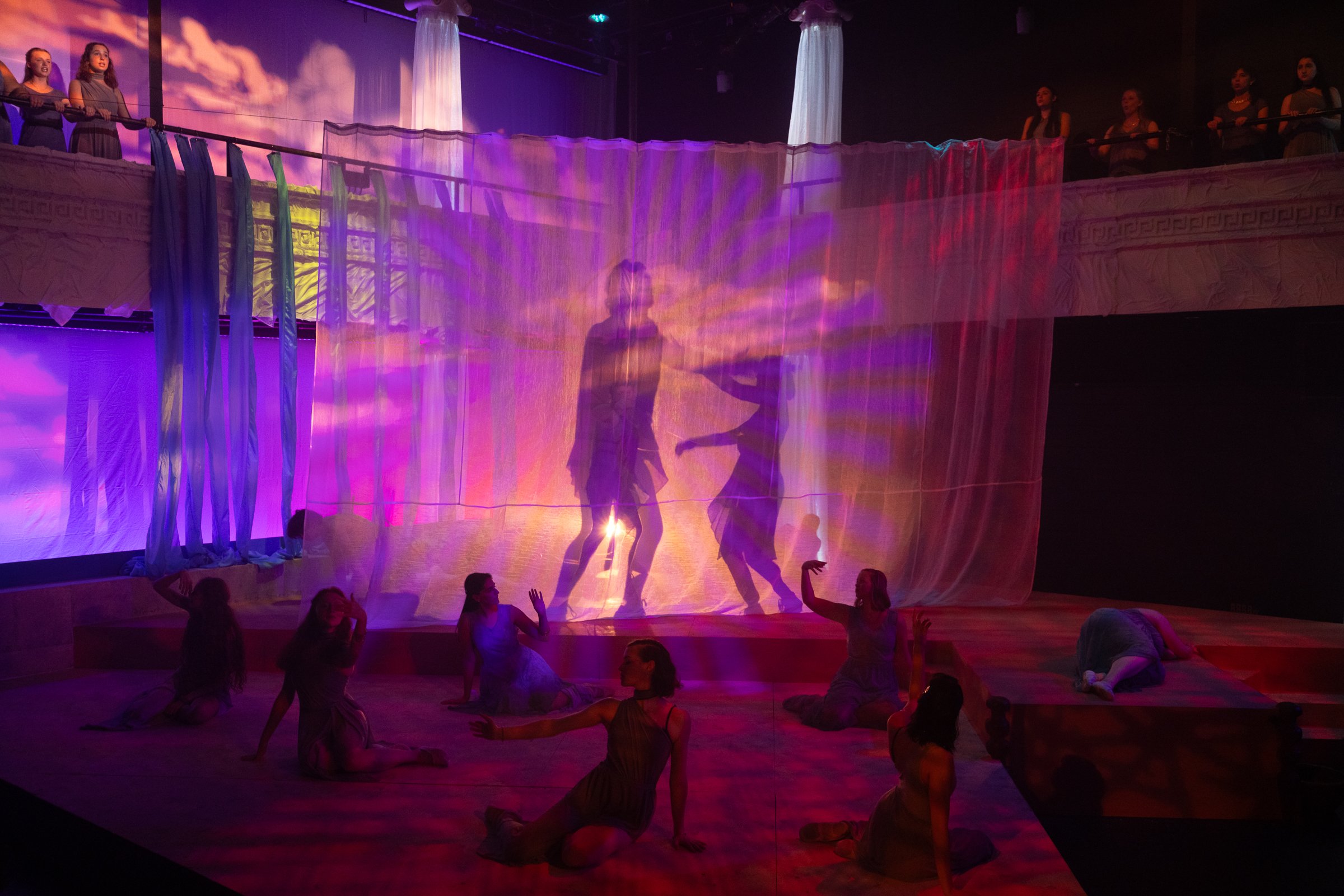 A stage performance with dancers, a colorful illuminated backdrop, and a curtain with a silhouette of a dancer projected on it, with people watching from the upper balcony.
