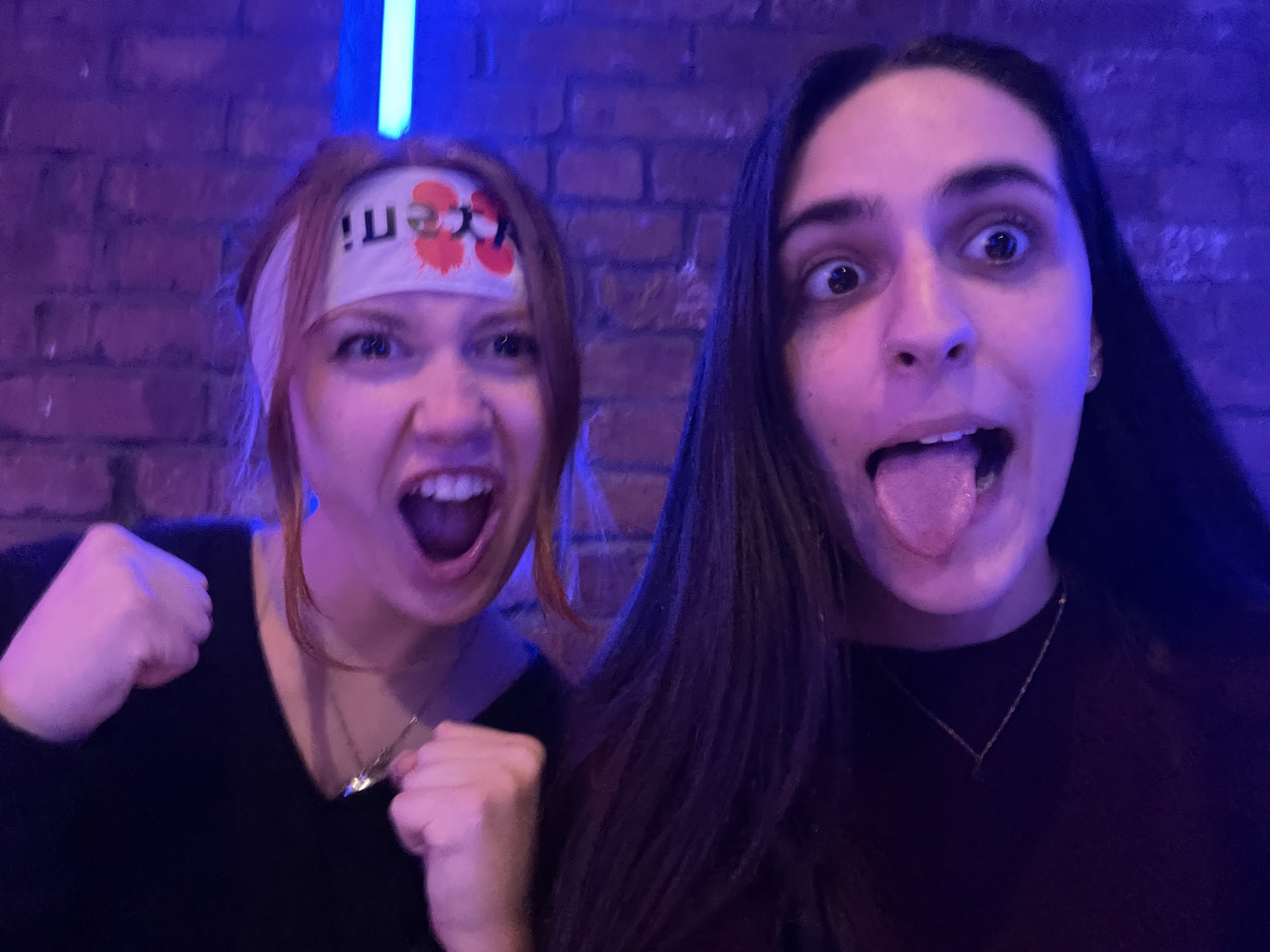 Two women making silly faces in a dimly lit, purple-hued indoor setting, with one wearing a hat or headband with the number 35 on it.