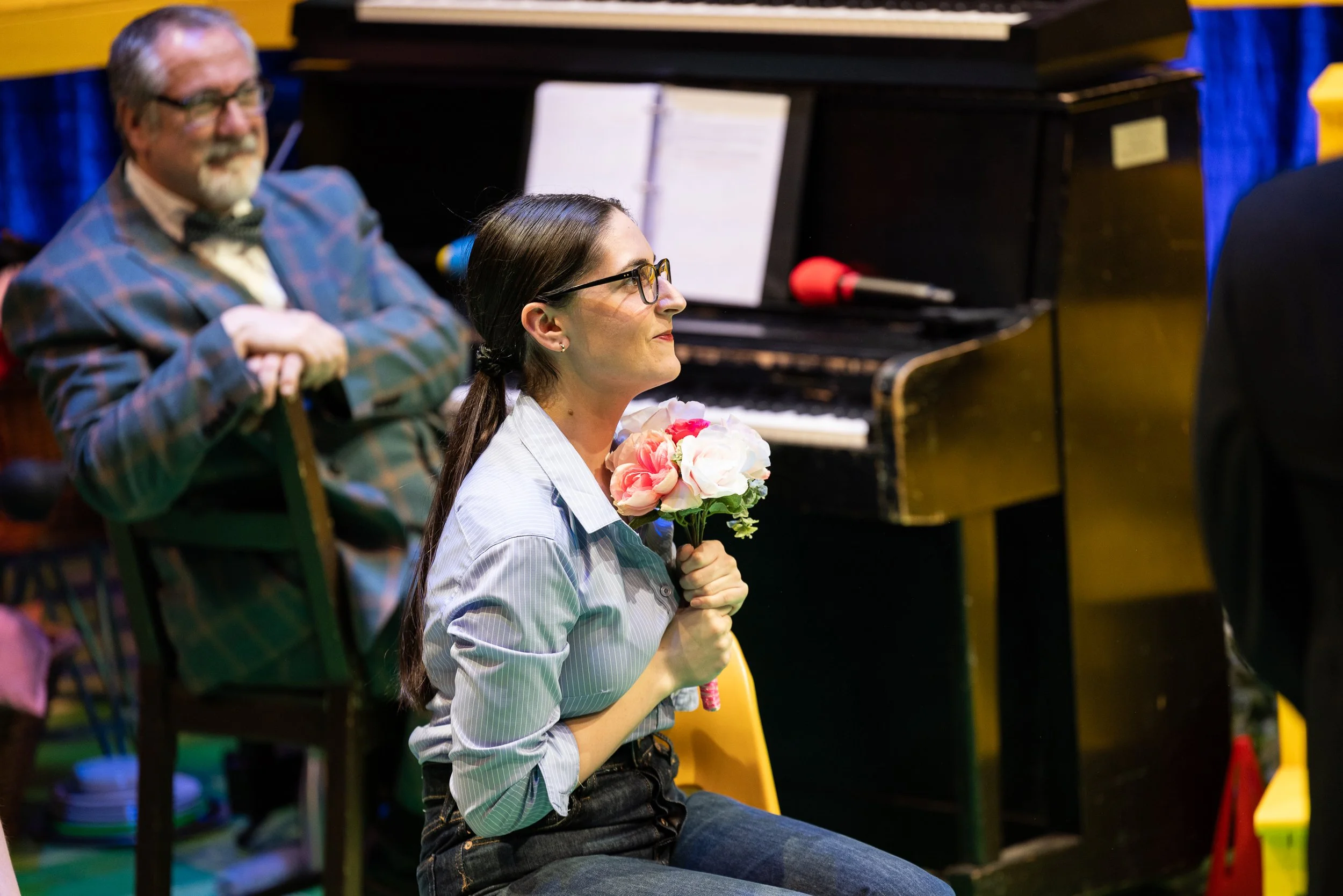 A woman sitting in a yellow chair holding a bouquet of pink and white flowers, with a man sitting behind her smiling, without glasses, in front of a black piano with sheet music and a red microphone.