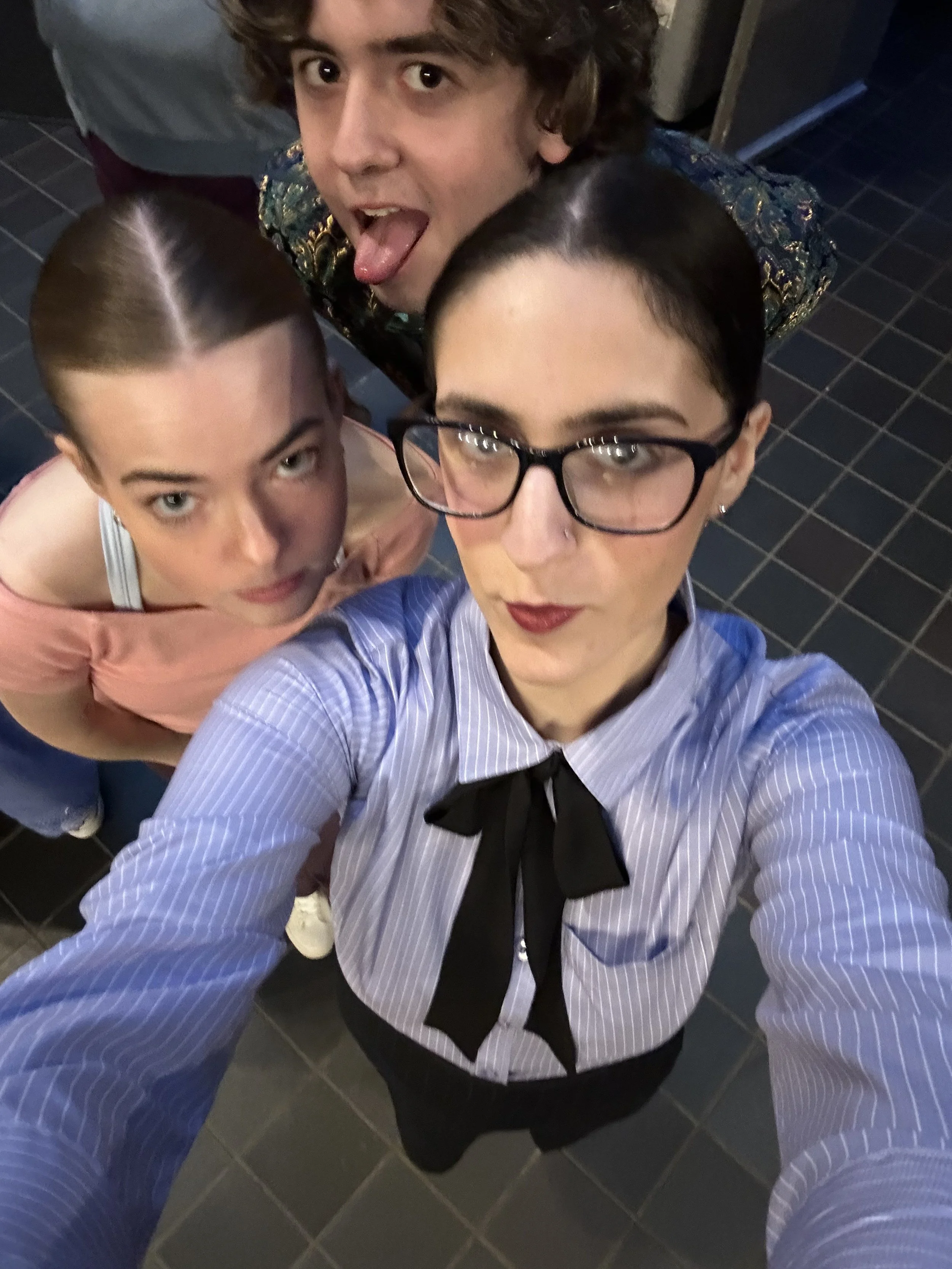 A selfie taken from above featuring three young women in a dimly lit environment with black tiled flooring. The woman in the foreground has dark hair, glasses, and red lipstick. The woman on the left has light brown hair in a braid, and the woman at 