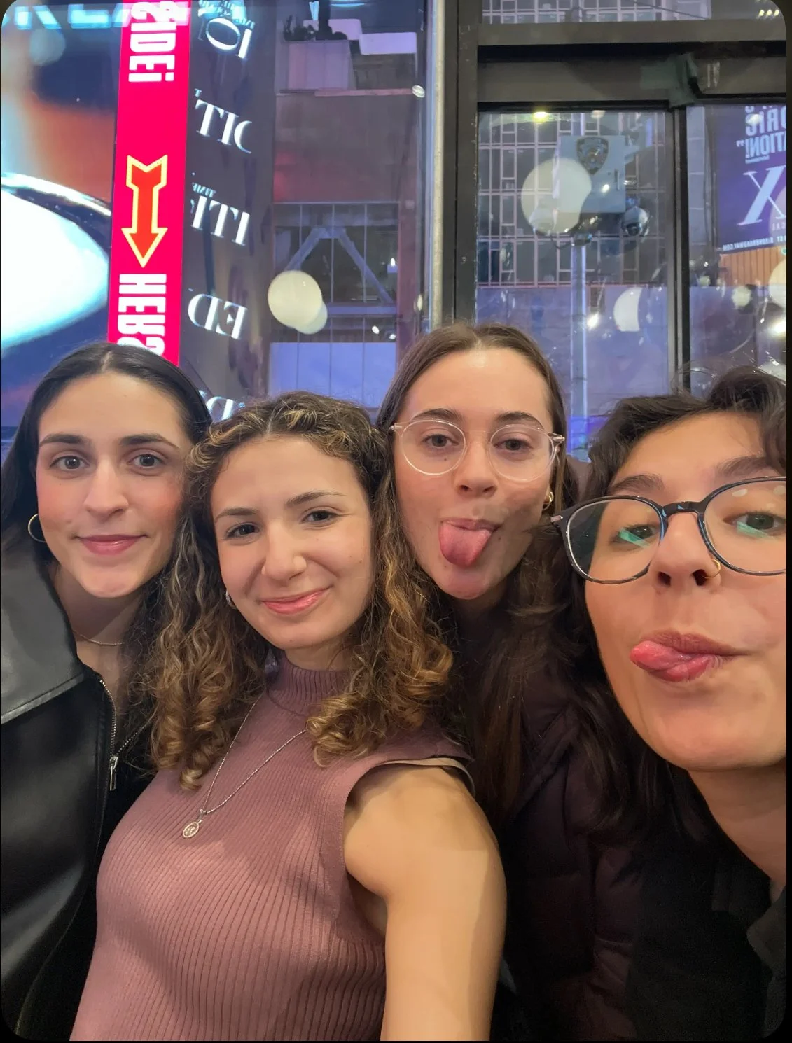 Four young women taking a selfie in a lively venue with bright lights and signs in the background. They are making playful faces and smiling.