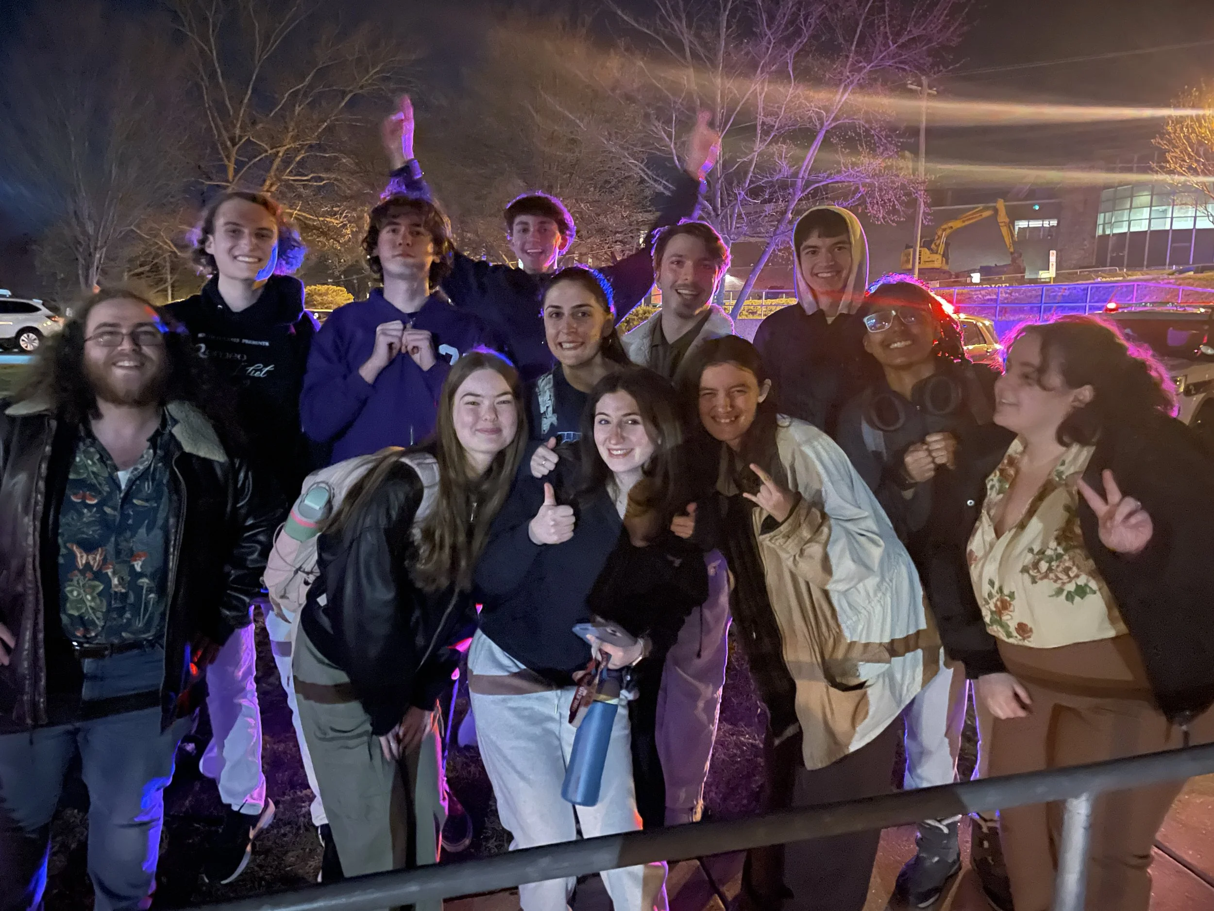 Group of young people enjoying nighttime outdoors with colorful lighting, some smiling, some making hand gestures, and a construction vehicle in the background.