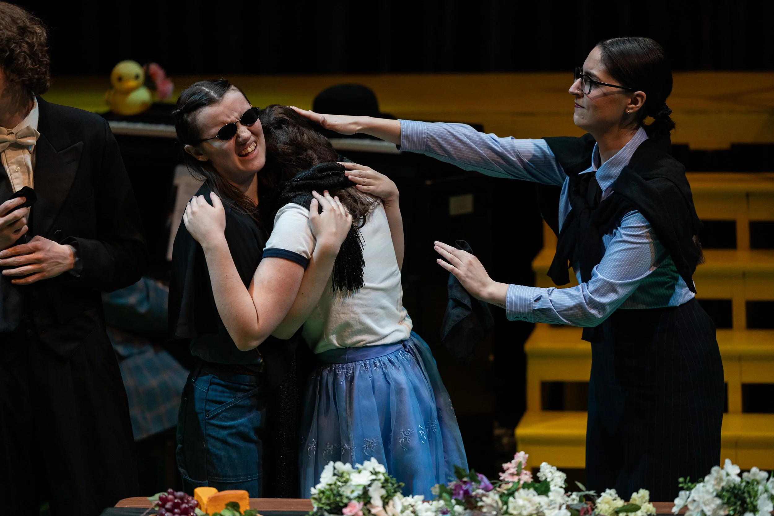 A woman with glasses and a blue skirt hugging a woman with dark hair and sunglasses at a celebration or wedding, with flowers and food in the foreground.