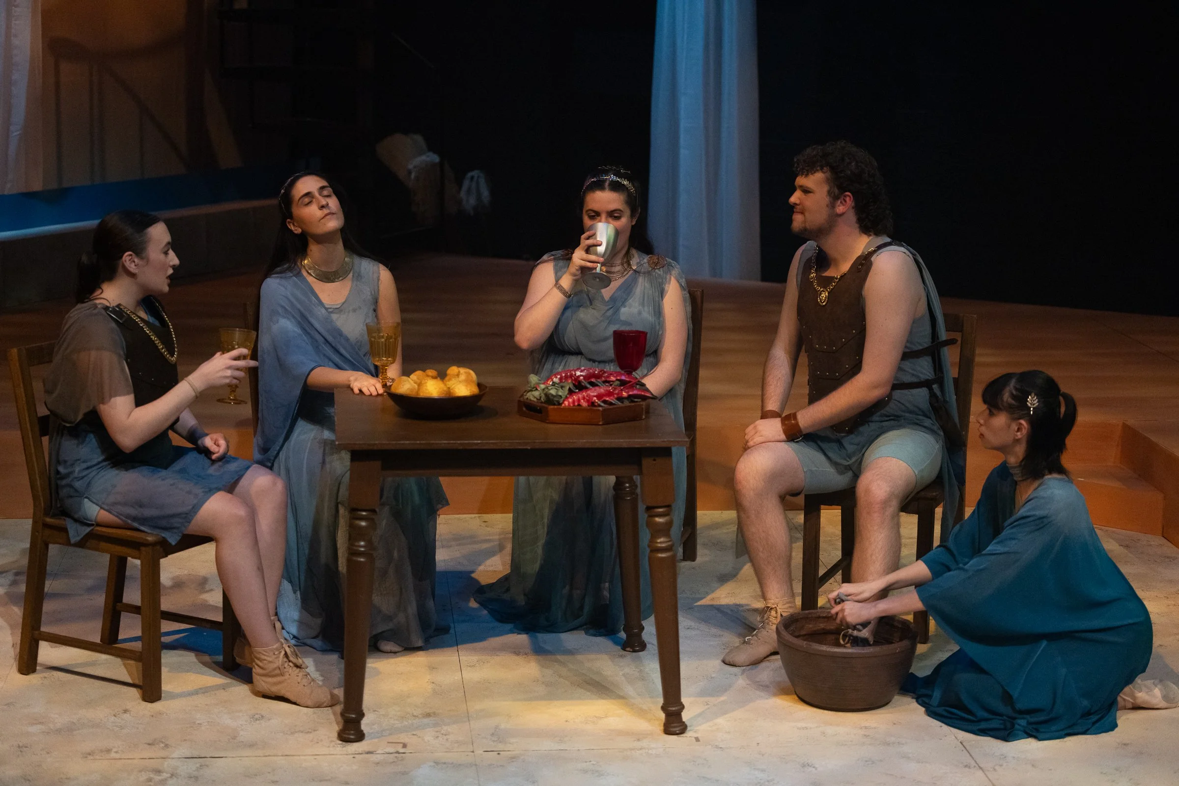 A theatrical stage scene with five actors dressed in toga-like costumes. One woman is drinking from a goblet, another woman has eyes closed and a third woman is seated on the floor. A man with curly hair and a beard is sitting at the table, and a you