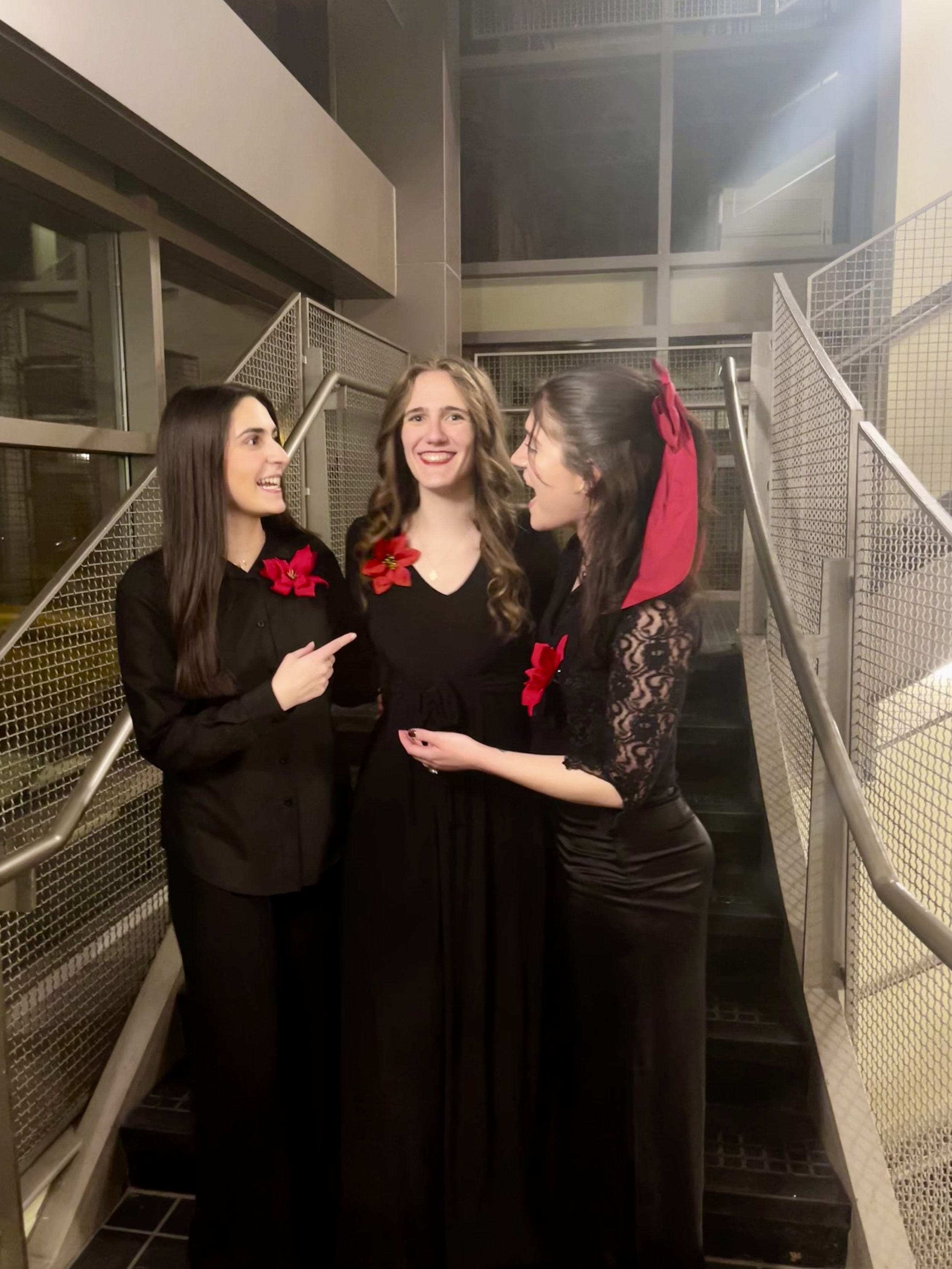 Three women dressed in black, with Christmas red flower accessories, celebrating and smiling together on a staircase in an indoor setting.