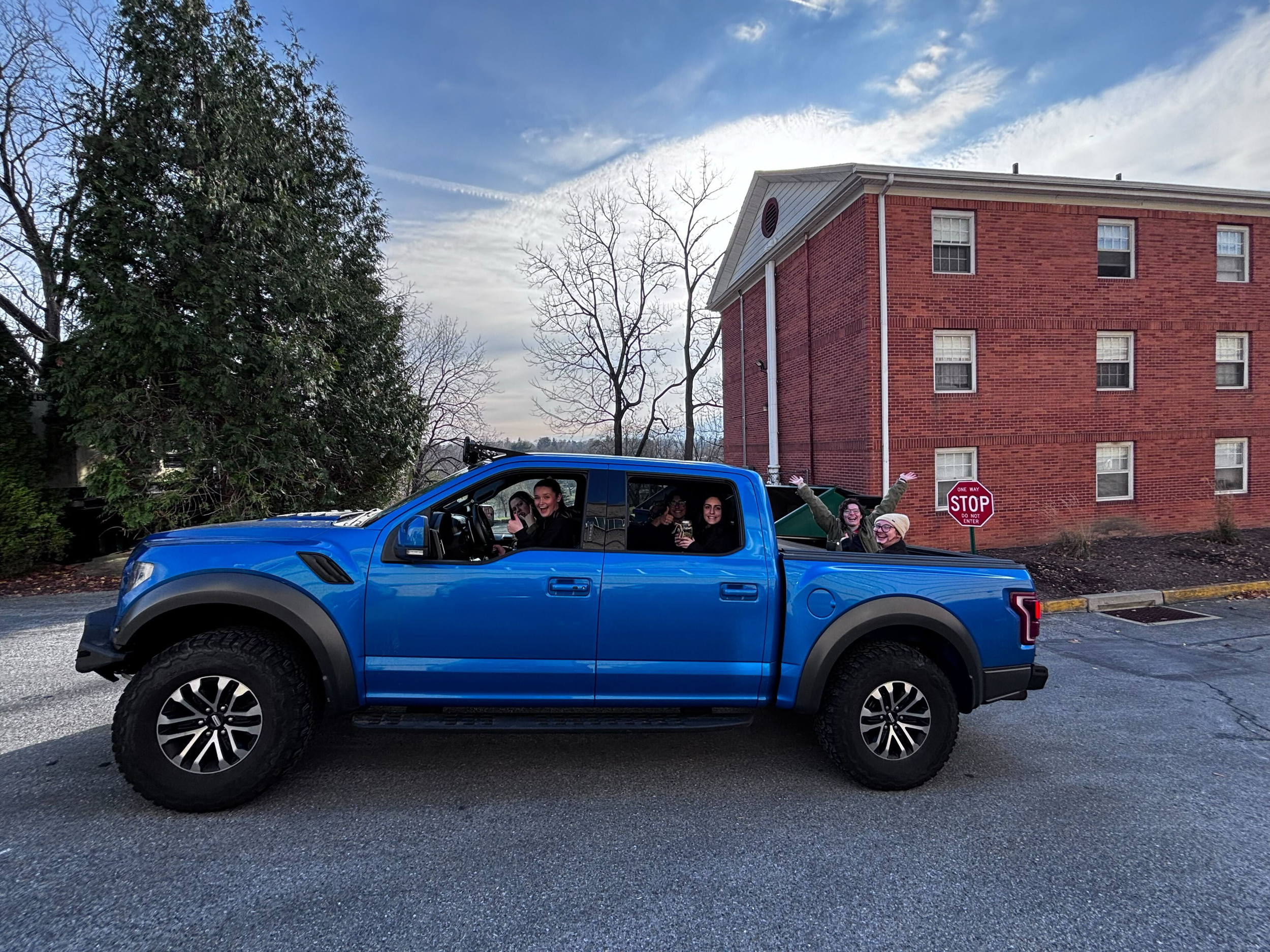 A blue pickup truck with five women inside, some smiling and waving, parked on a street in front of a red brick building, trees, and a stop sign.