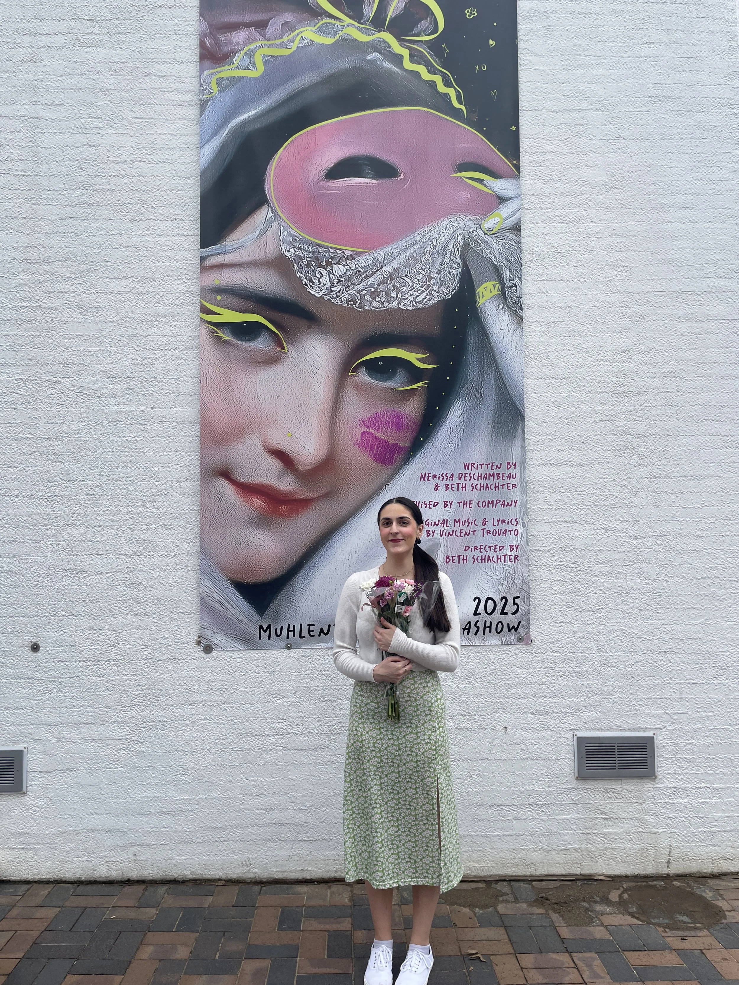 A young woman with dark hair in a ponytail wearing a white long-sleeve shirt, a green patterned skirt, white sneakers, and holding a bouquet of flowers, stands in front of a large colorful artistic mural of a woman's face with makeup and abstract ele