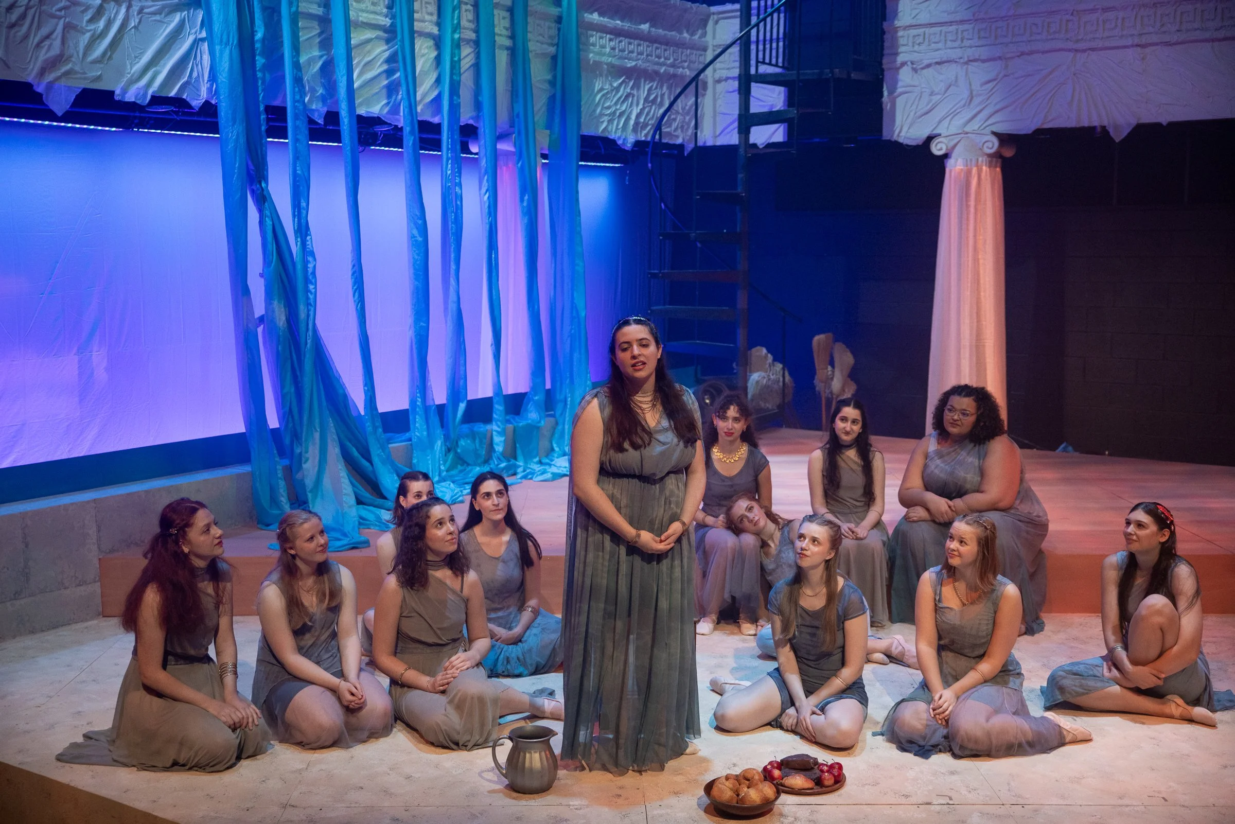 A group of women and girls dressed in ancient Greek-style costumes, sitting and standing on stage with a blue and white backdrop, while one woman in the foreground appears to be performing or speaking.