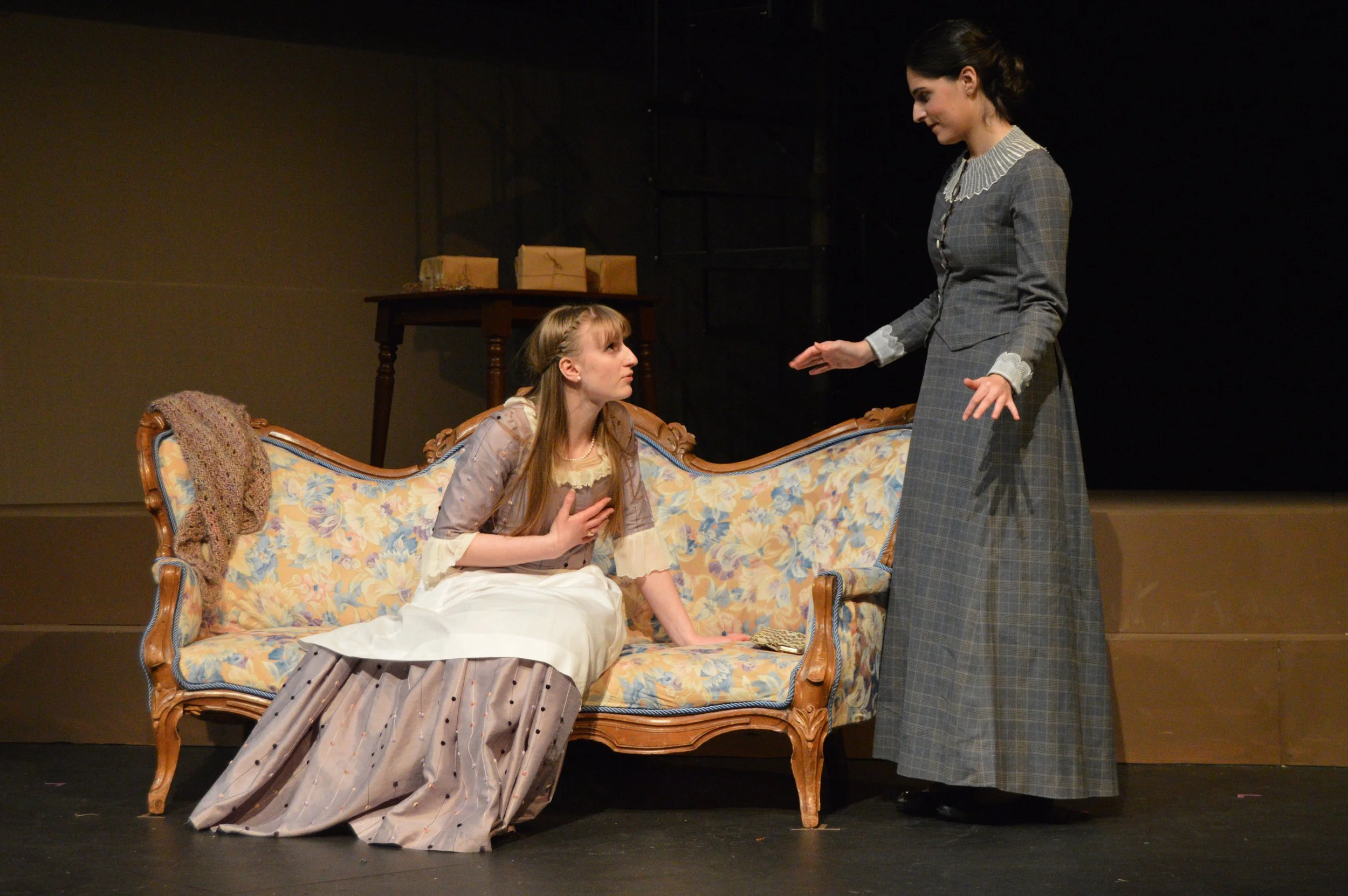 Two women in vintage dresses engaging in a dramatic scene on stage. One woman with blonde hair is sitting on a floral-patterned sofa, holding her chest and looking up with a concerned expression. The other woman with dark hair, standing and dressed i