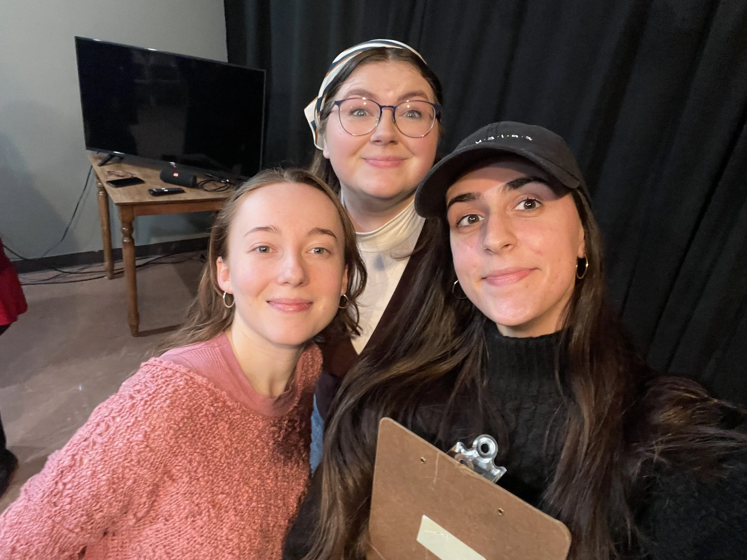 Three young women taking a selfie in a room with a black curtain background, with a television and wooden table behind them.