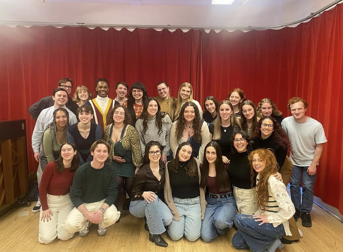 A group of young people standing and sitting in front of red curtains, posing for a group photo indoors.