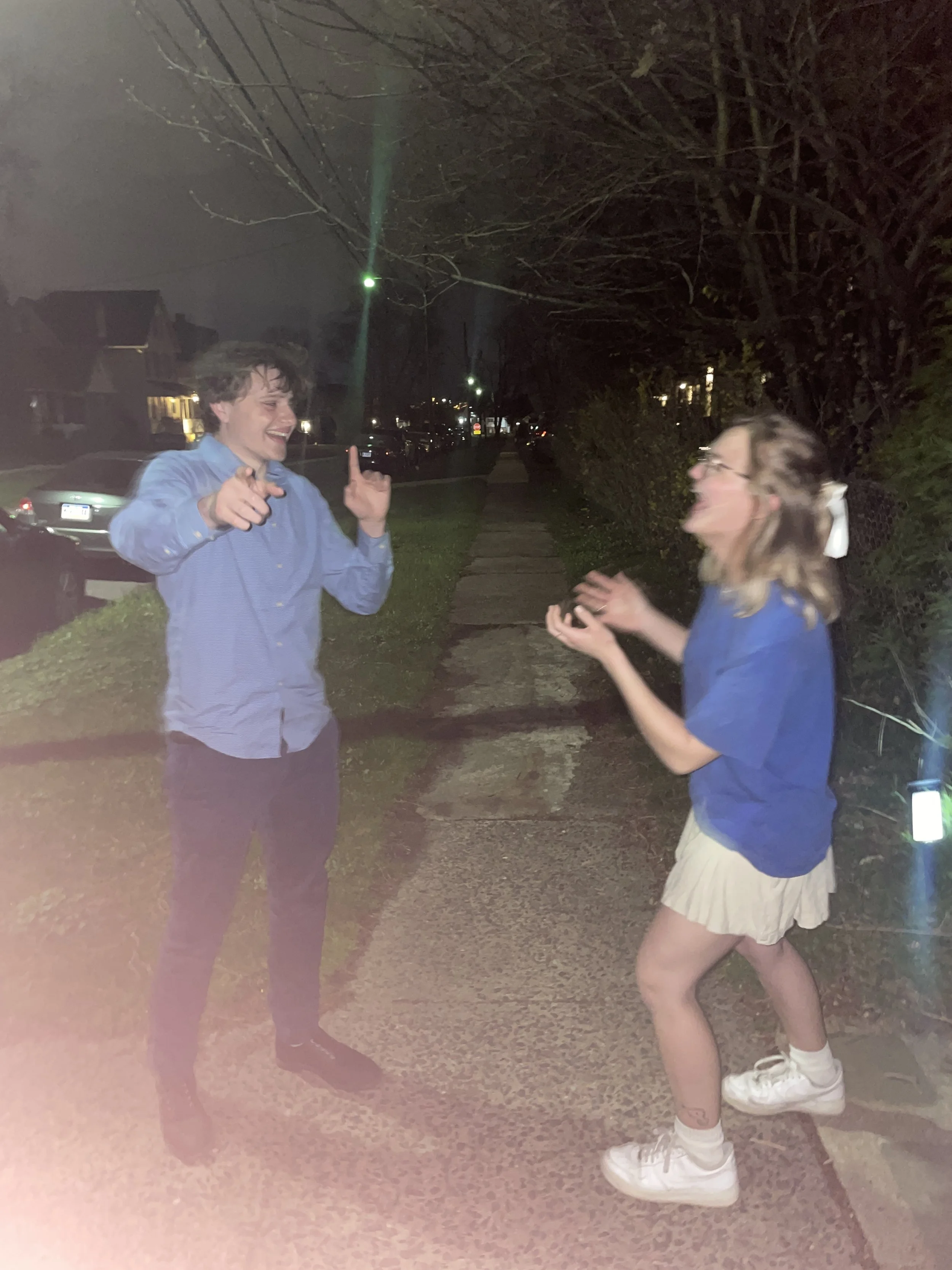 Two people, a young man and a young woman, are standing on a sidewalk at night, engaging in a lively conversation. The young man is pointing and smiling, while the young woman is gesturing with her hands, with her mouth open in an animated expression