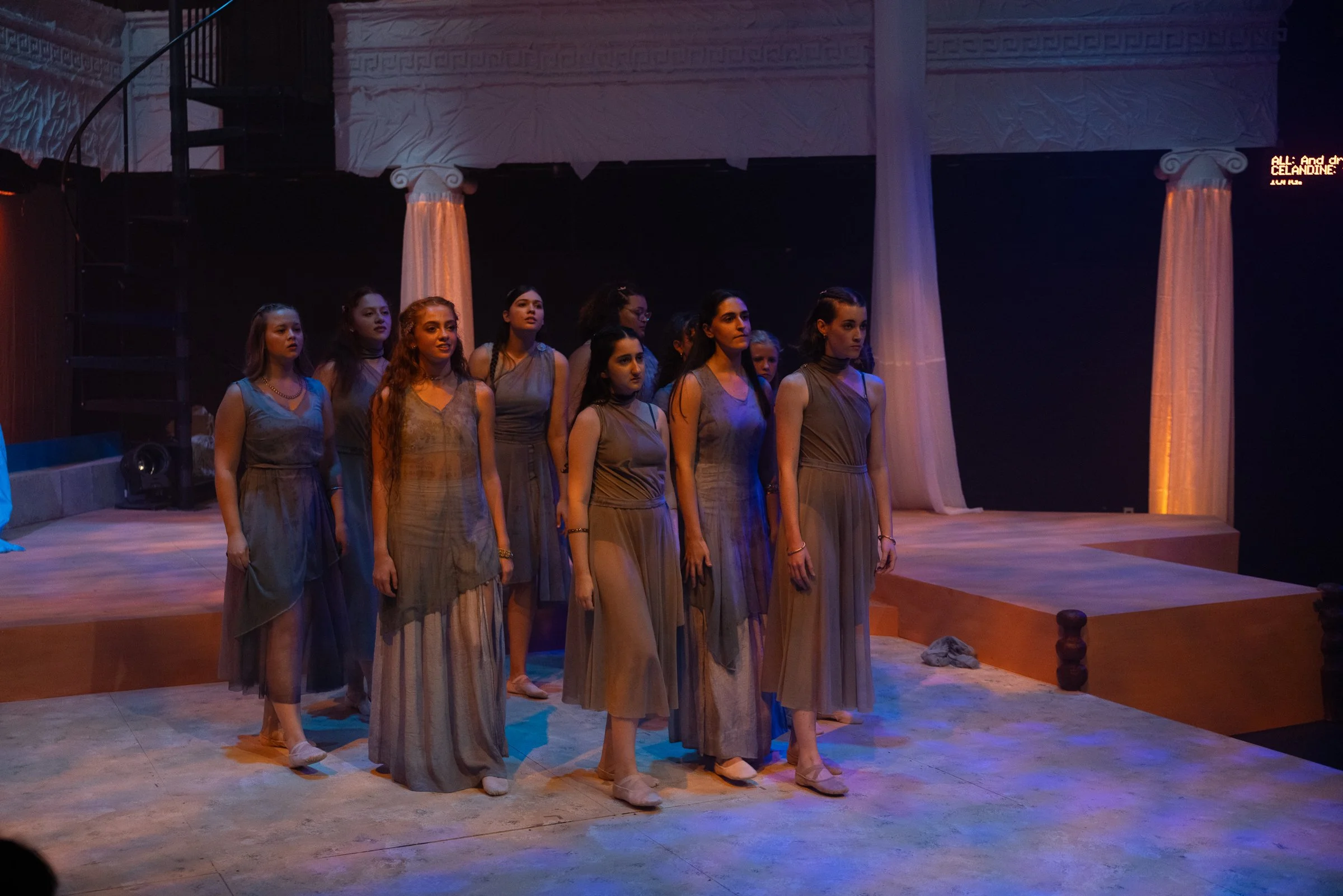 A group of young women dressed in matching grey costumes standing on a concert stage, facing forward with solemn expressions, with Greek-style columns and draped fabric in the background.
