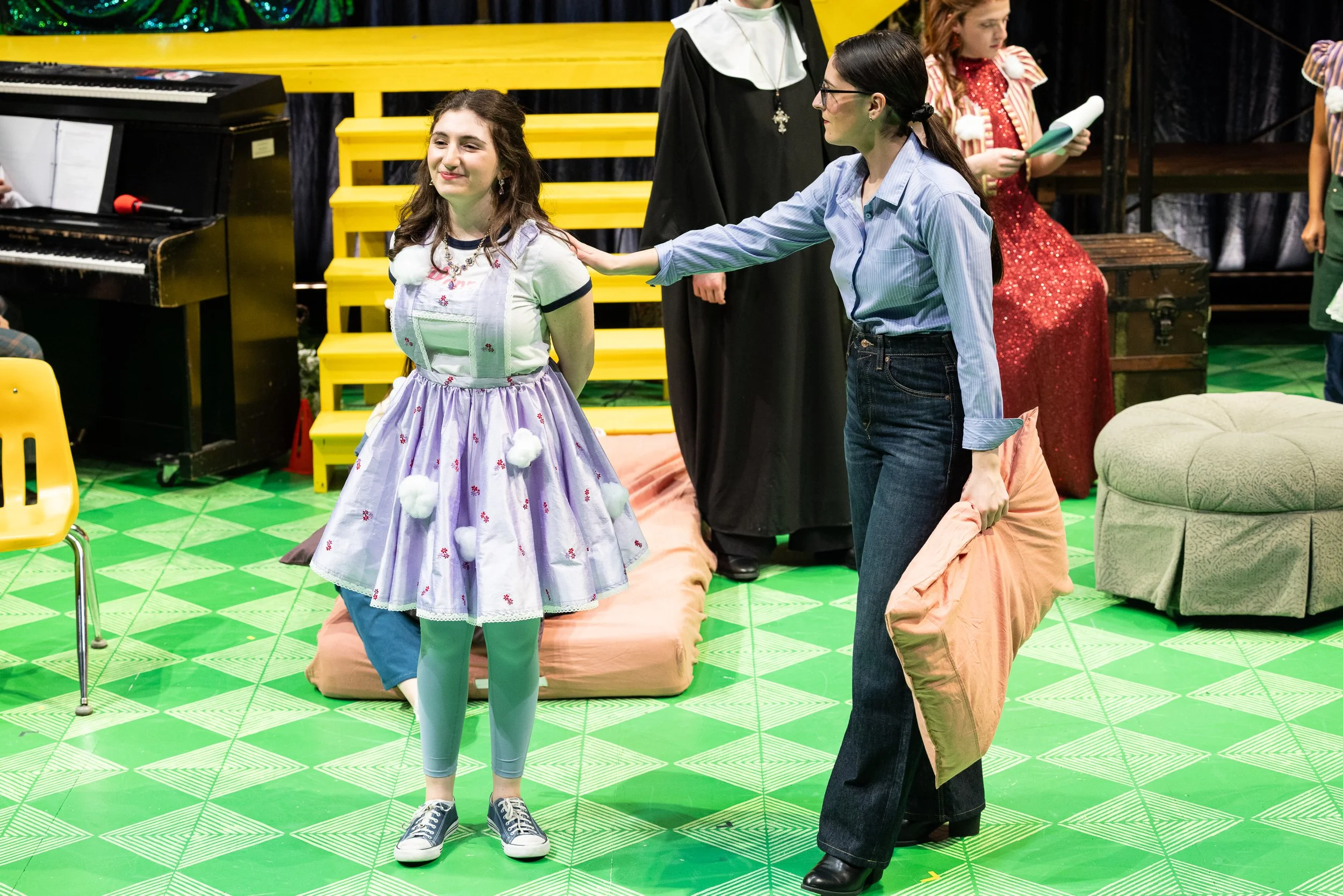 Two women are on stage with theatrical props, one is wearing a pink and purple costume, and the other is dressed in casual clothes, touching the first woman's shoulder. In the background, there are children, a keyboard, and colorful set decorations.