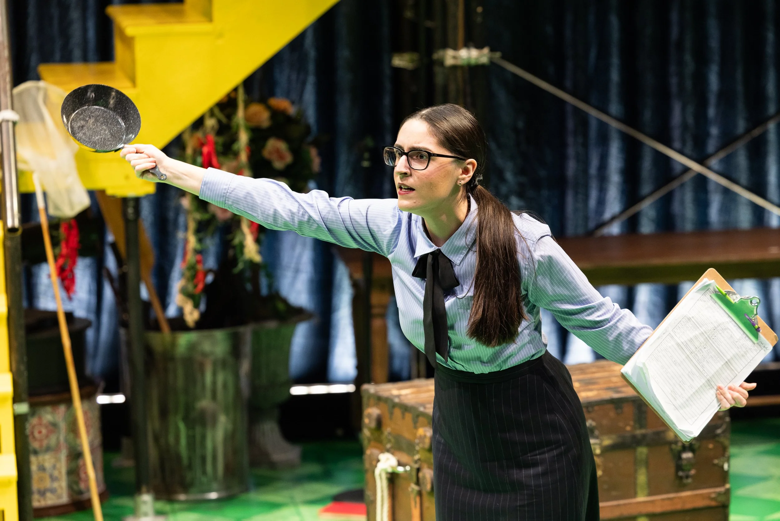 Woman in glasses with dark hair, wearing a striped shirt and black skirt, raising a black pan in a room with dark blue curtains and colorful decorations.