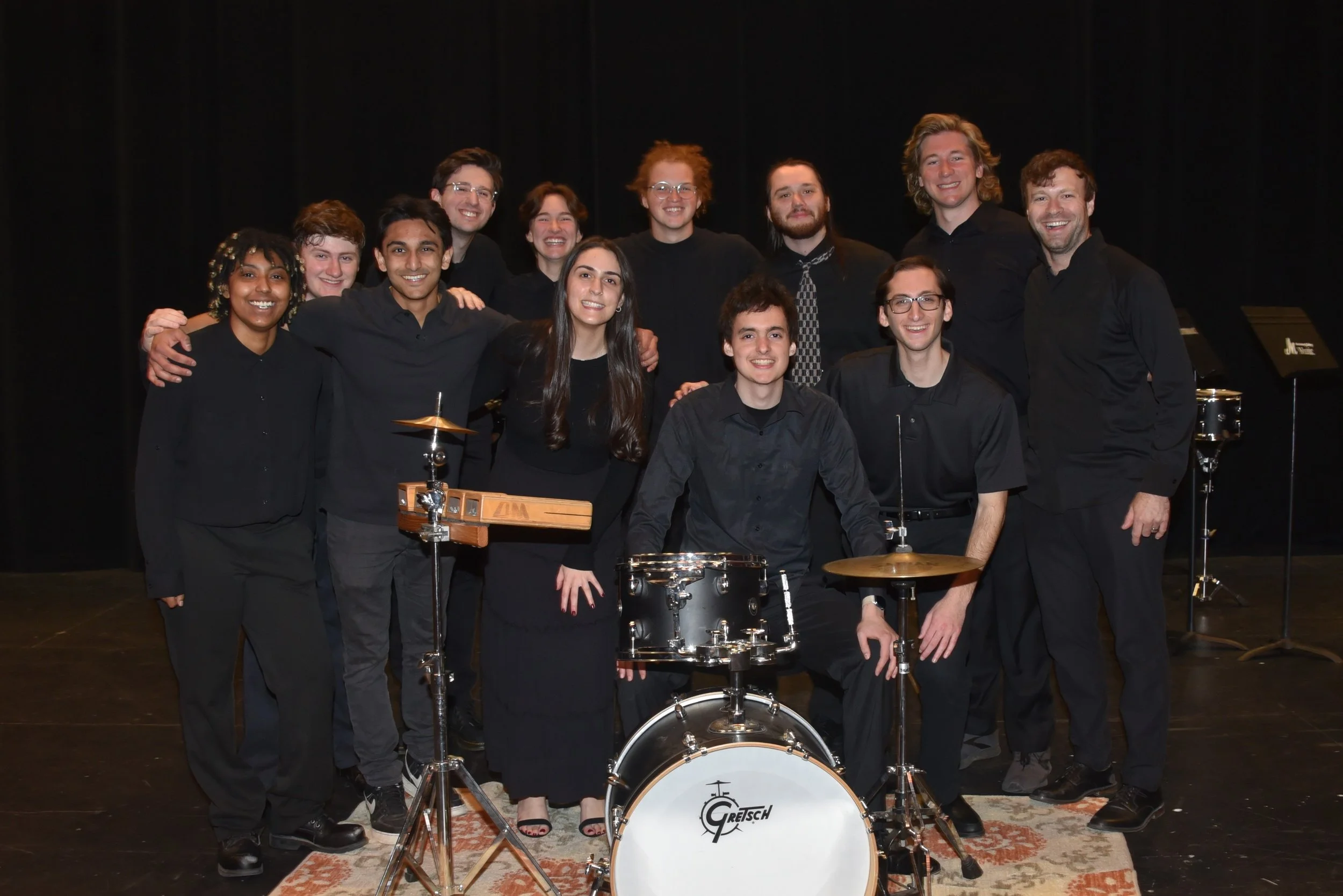 Group of eleven people dressed in black, standing on stage with musical instruments, including drums and a music keyboard, in a dark setting.