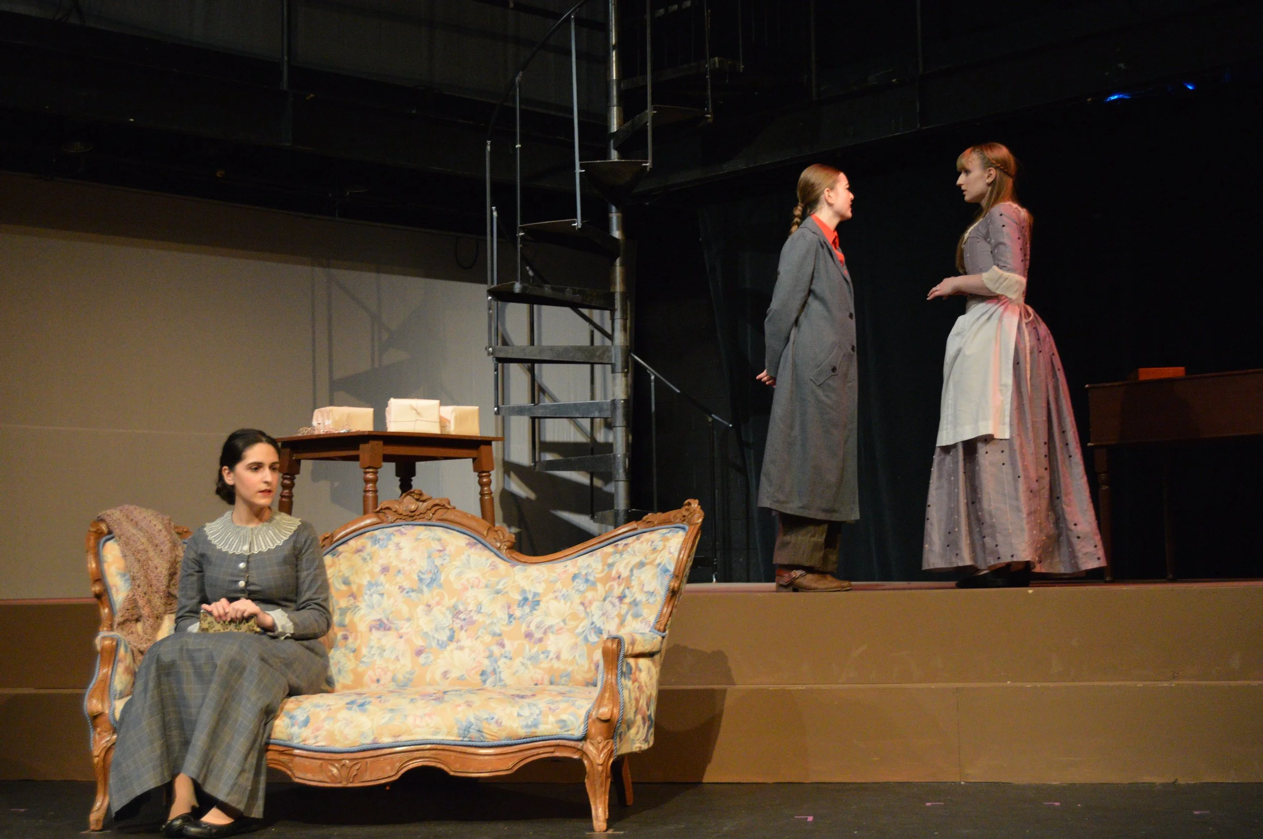 The image shows a stage scene from a play with four women in period costumes. Two women are standing and facing each other in the center of the stage, and two women are sitting on a vintage floral sofa and a wooden chair to the side. The stage has mi