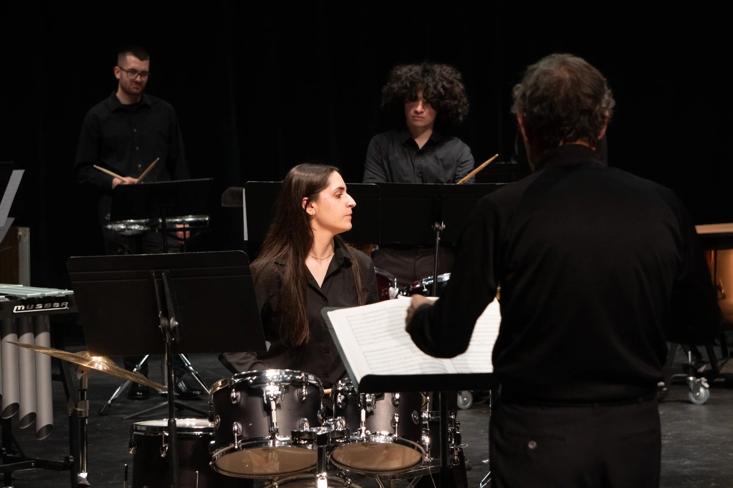 A group of musicians on stage, with a woman playing drums and a man conducting, during a performance.