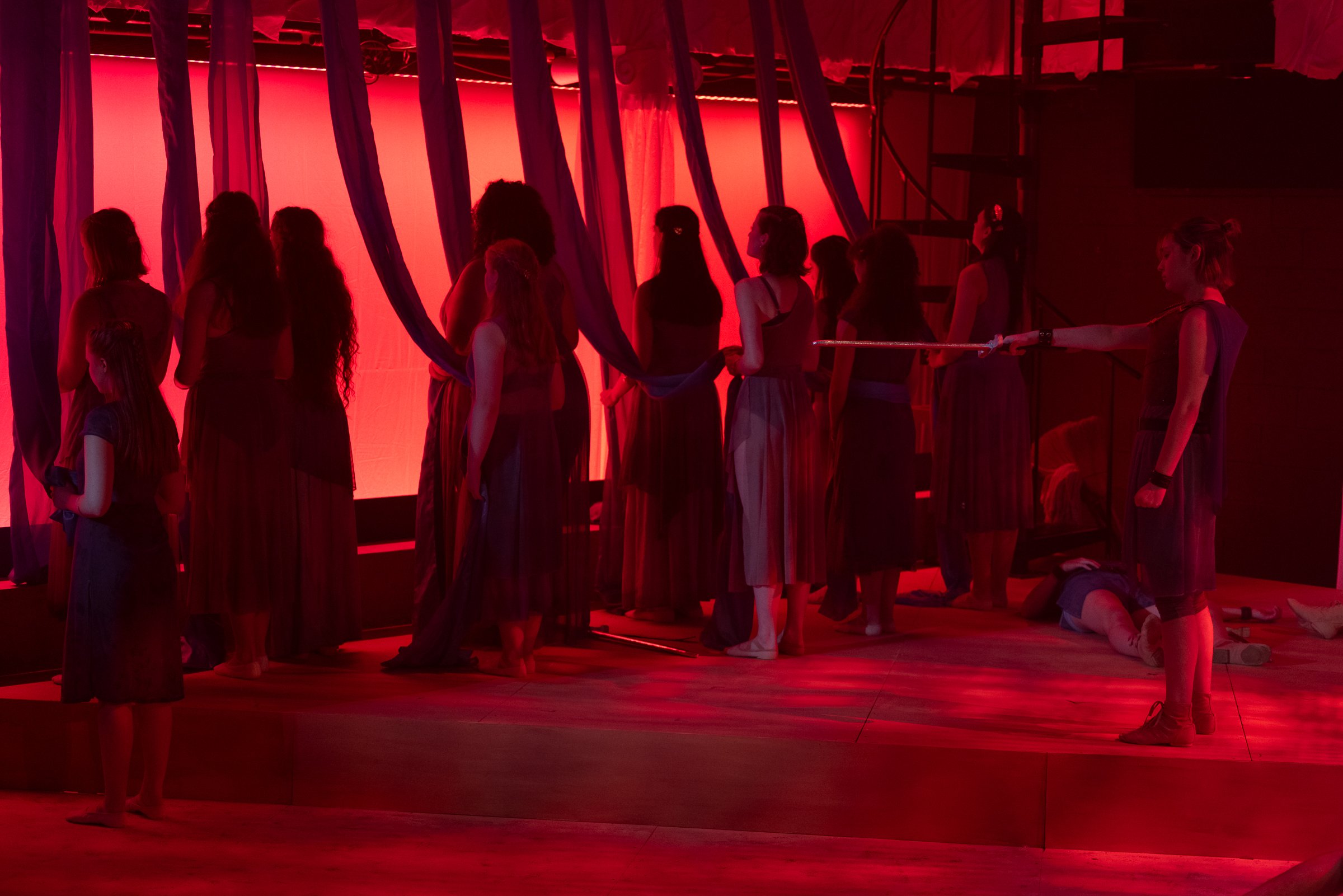 Multiple women dressed in red and black costumes in a dimly lit room with red lighting and fabric drapes, possibly performing or rehearsing.