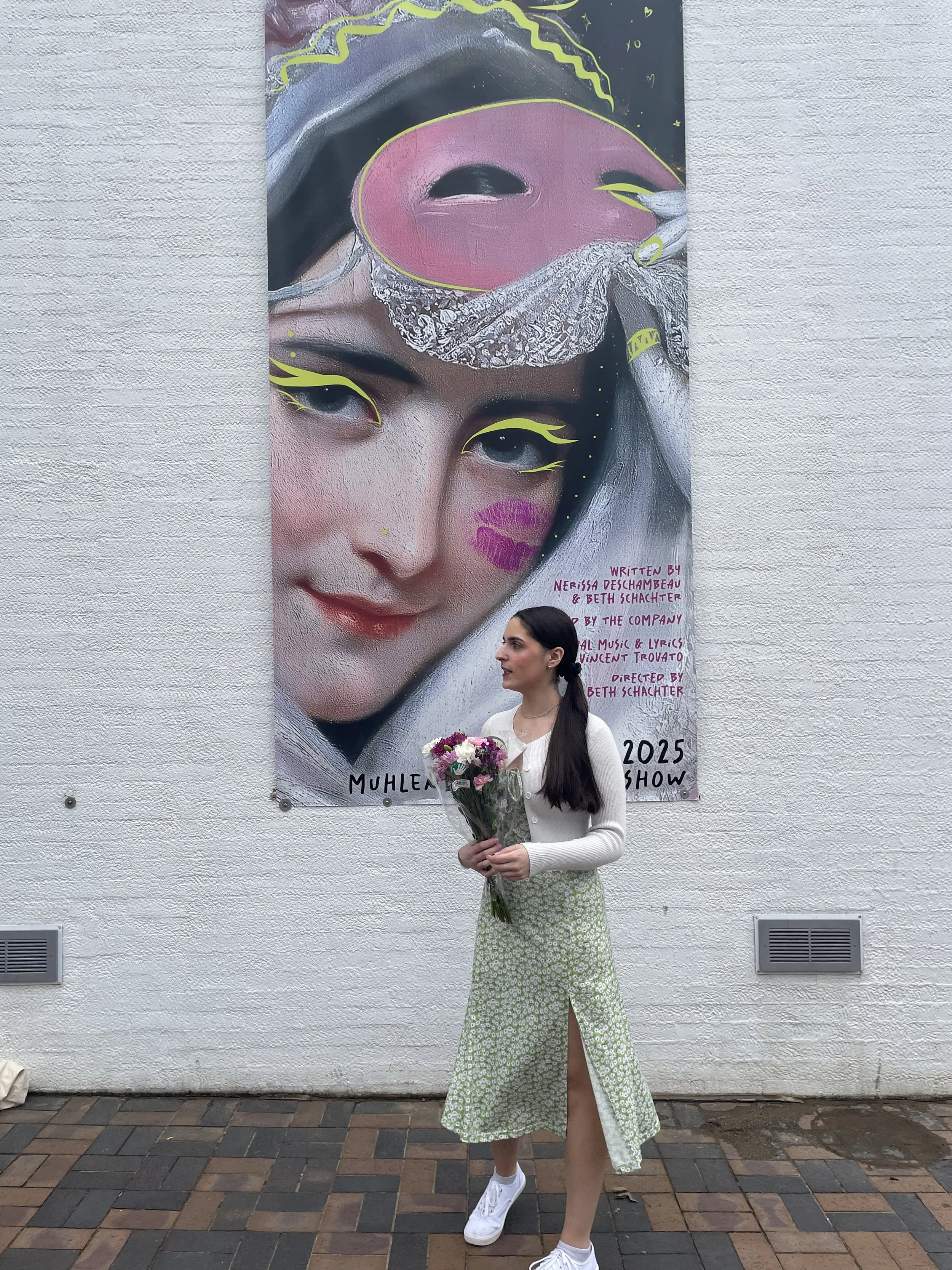 A young woman standing in front of a large colorful poster of a female face with artistic makeup and bright accents, holding a bouquet of flowers, wearing a white top, a green patterned skirt, and white sneakers.