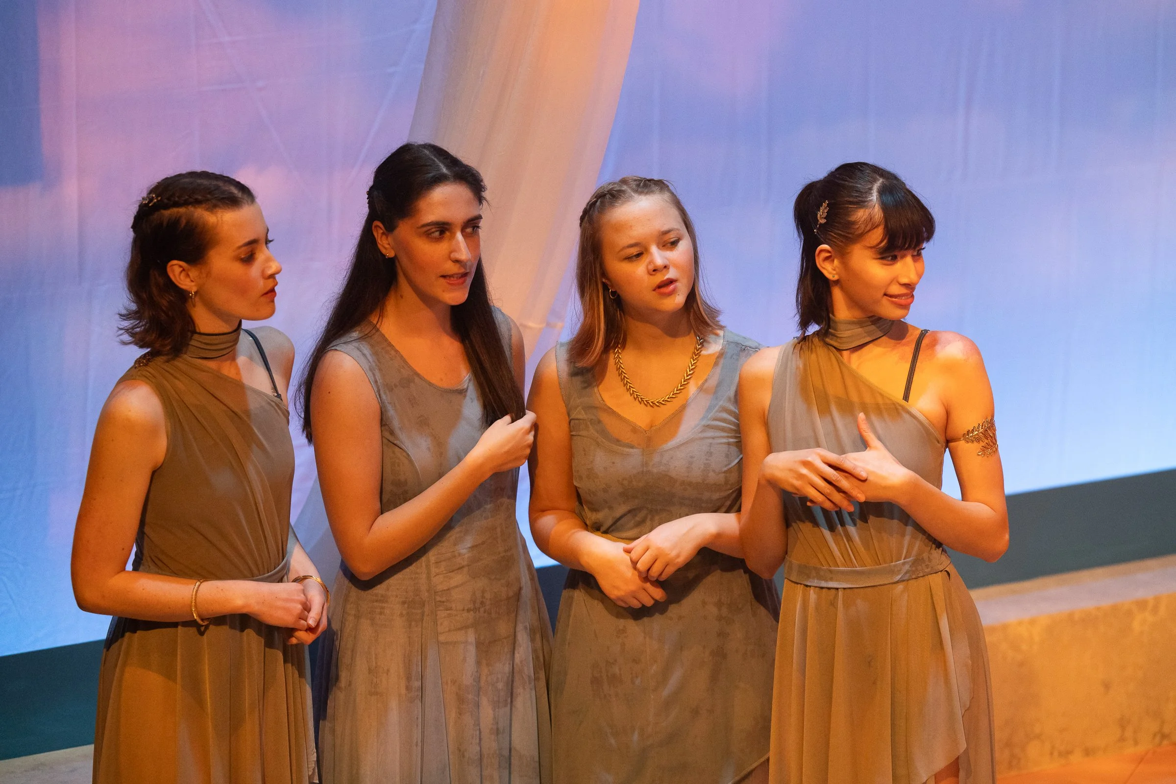 Four women standing together in elegant dresses, engaging in conversation at an indoor event with a purple and blue backdrop.