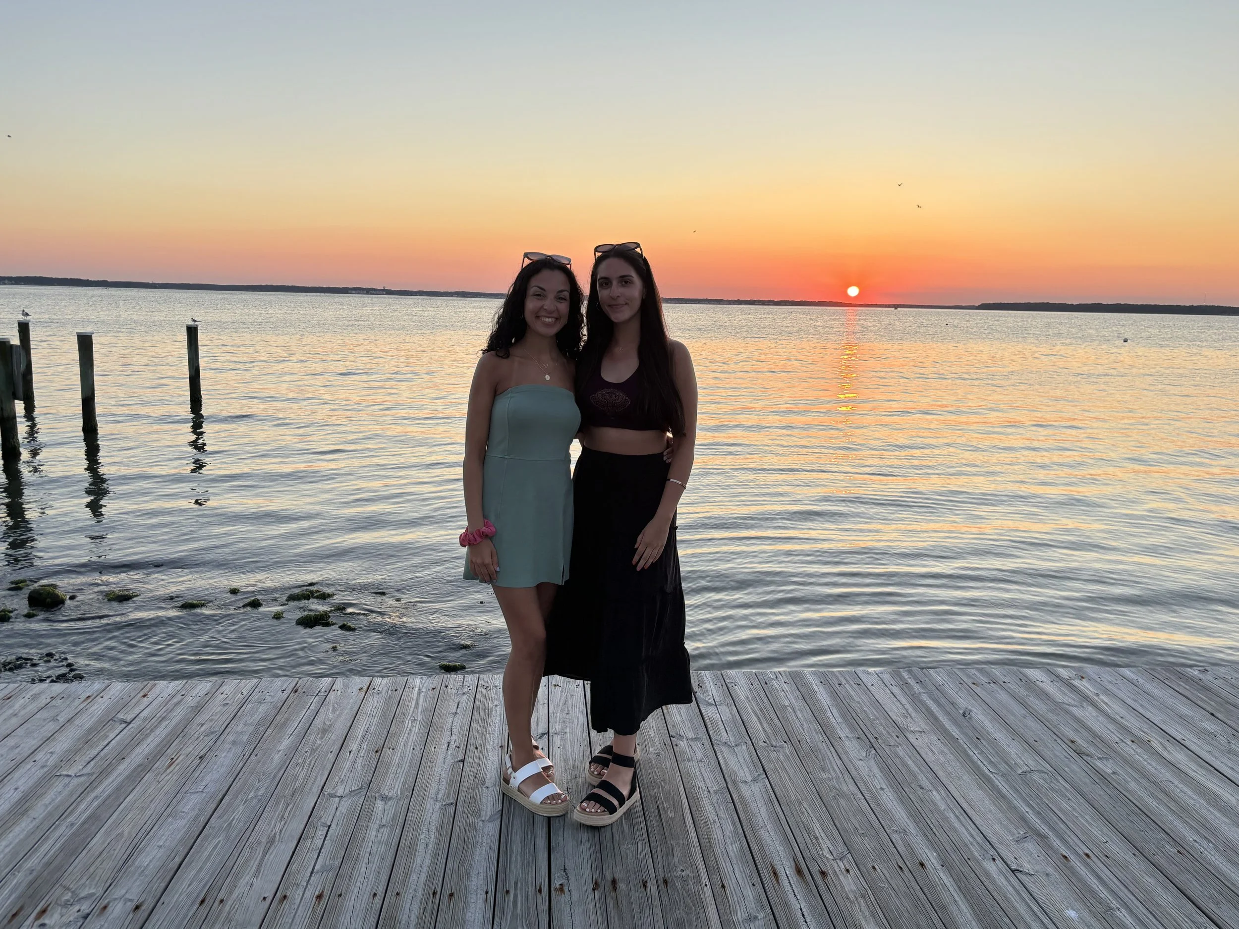 Two young women standing on a wooden dock by the water during sunset. The woman on the left wears a light blue strapless dress, and the woman on the right wears a black crop top and black skirt. Both have sunglasses on their heads and are smiling at 