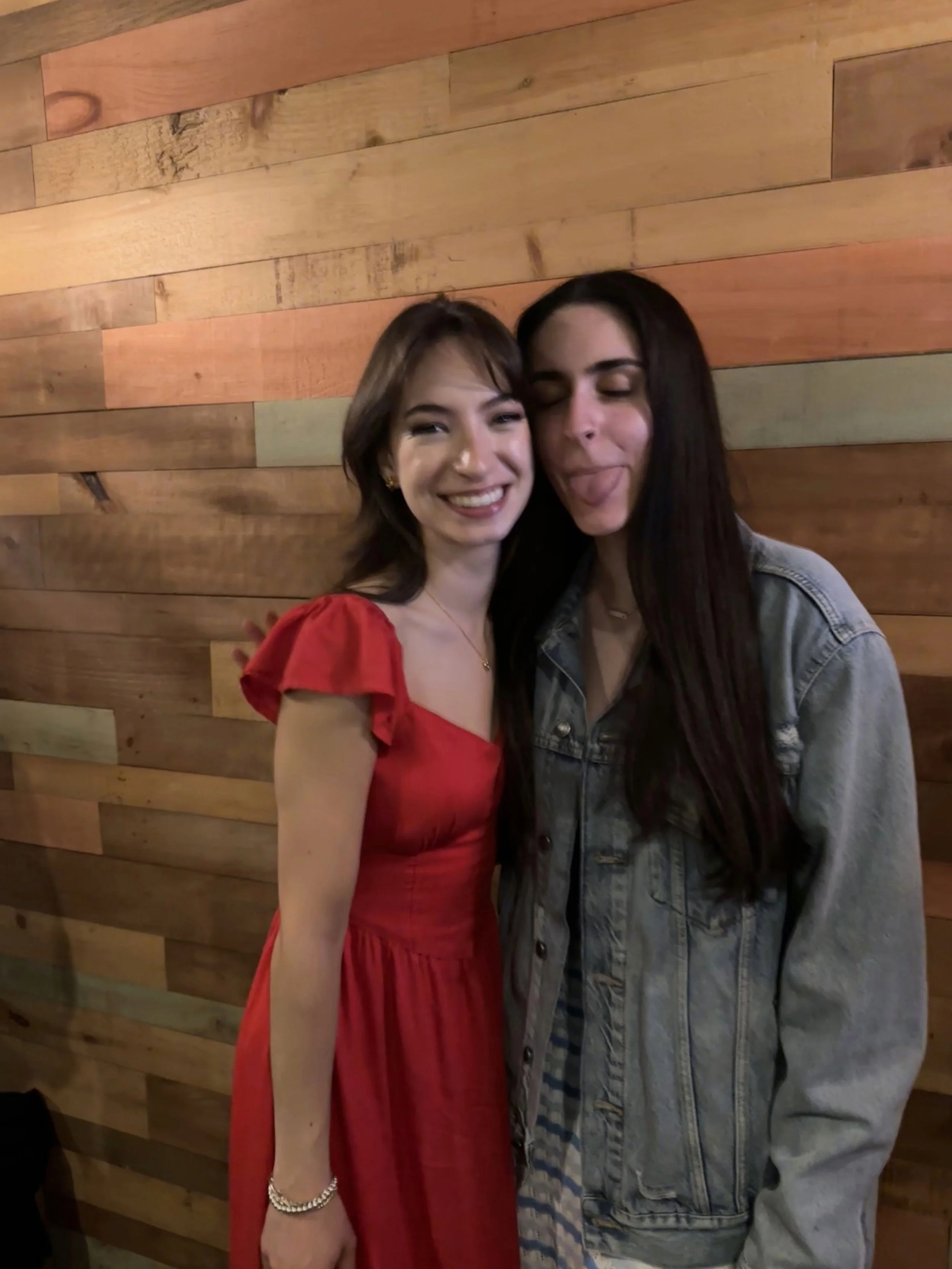 Two women posing with one sticking out her tongue against a wooden wall background.