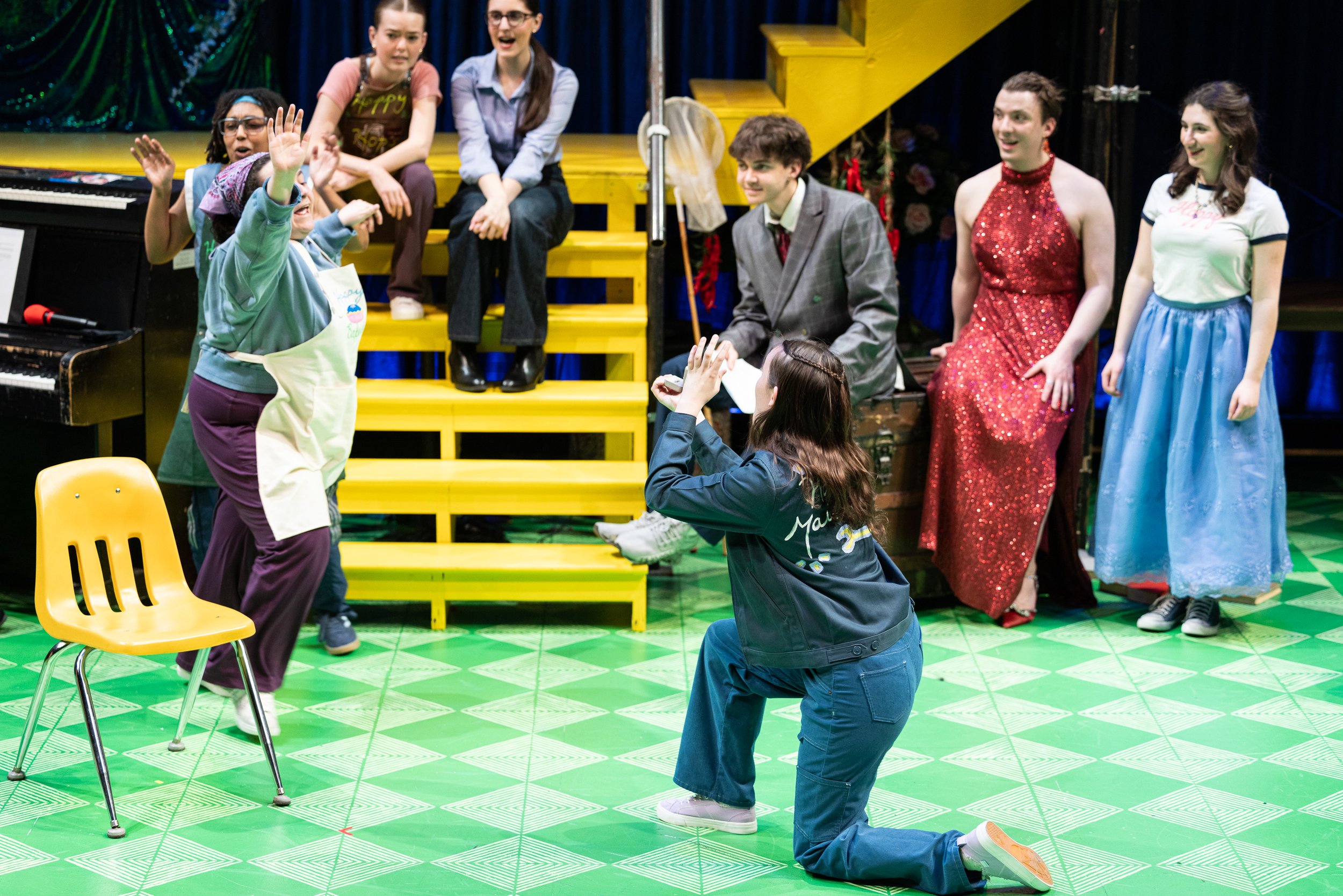 People performing a comedy skit on a stage with yellow steps, some seated and some standing, in front of a blue curtain with Christmas decorations.