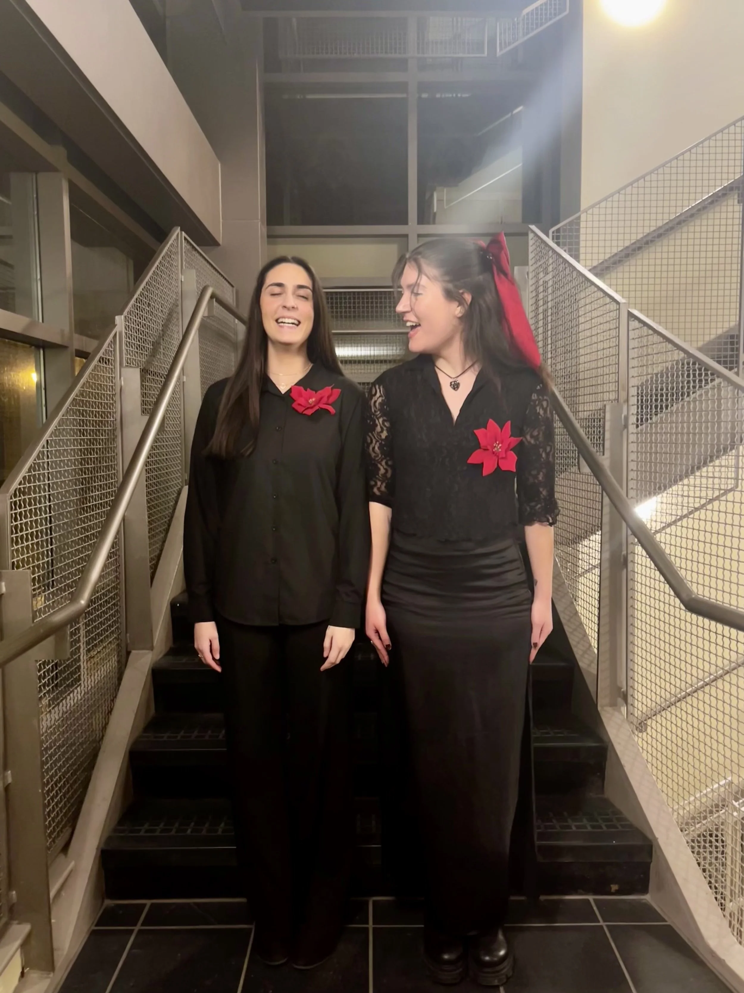 Two women standing on a staircase, smiling and laughing, wearing black clothing with red poinsettia decorations pinned to their shirts.