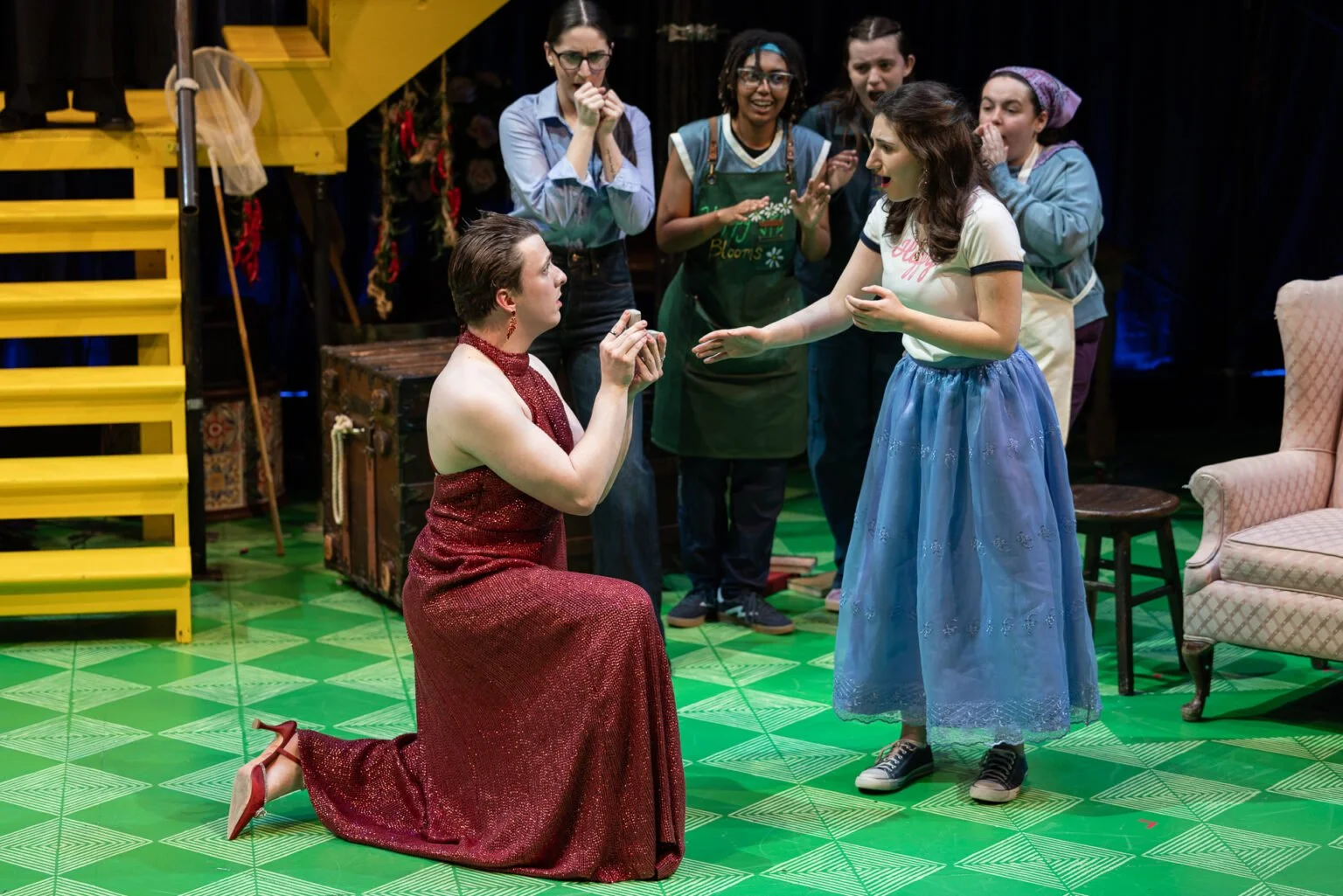 A theatrical scene with a woman dressed in a red, sparkly gown kneeling on the green patterned floor and looking at a girl in a light blue skirt and sneakers, as if about to ask her a question or make a plea. The girl is reaching out to the woman, ap