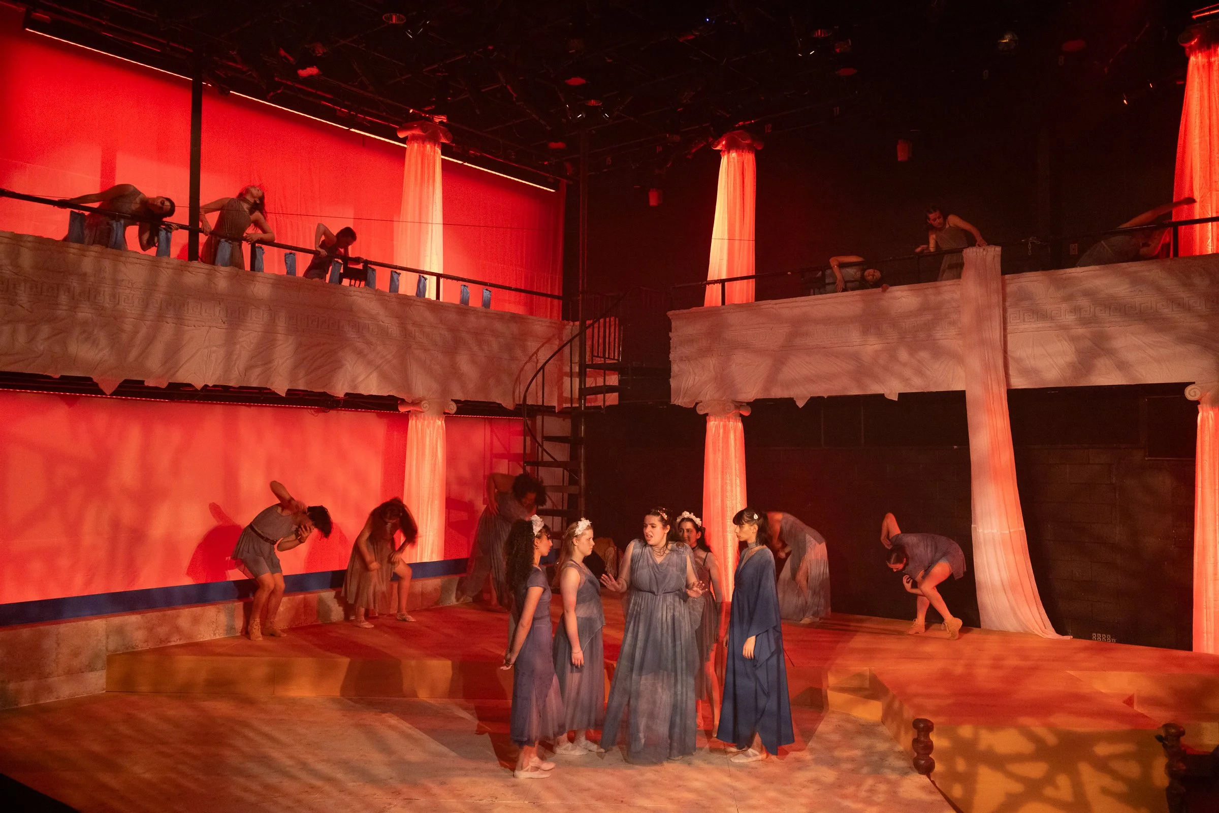 The image shows a stage scene with performers dressed in Greek-style costumes, standing and kneeling on a wooden stage with a two-level set. The set features red fabric curtains and columns, with a red backdrop illuminated by stage lighting.