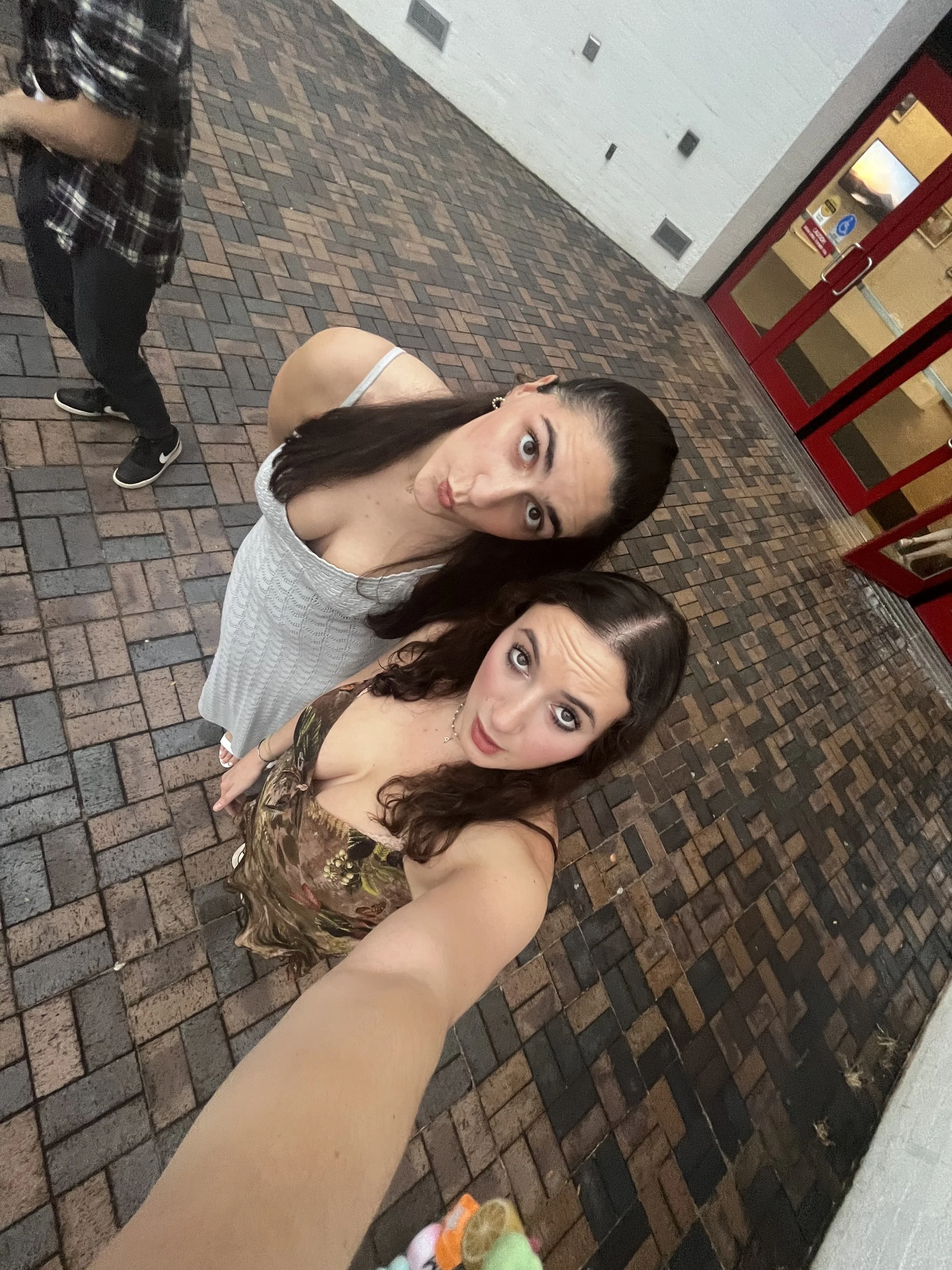 Two women taking a selfie outside at night, with a brick sidewalk and a red door in the background. One woman is wearing a patterned dress, and the other is in a tank top. A person wearing a plaid shirt and sneakers is partially visible on the left s