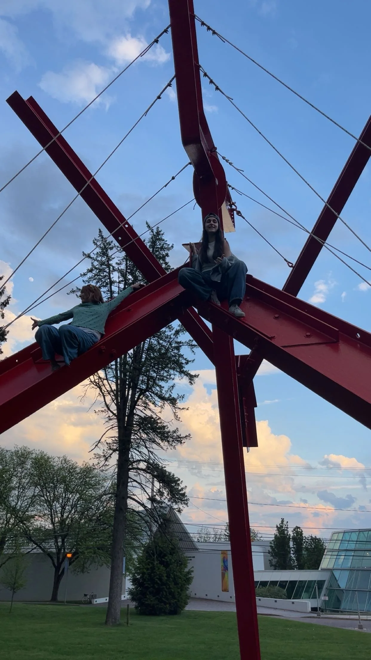 A playground structure with two children sitting on it, one in a blue shirt and the other in a gray shirt, against a backdrop of trees, a modern building, and a partly cloudy sky.