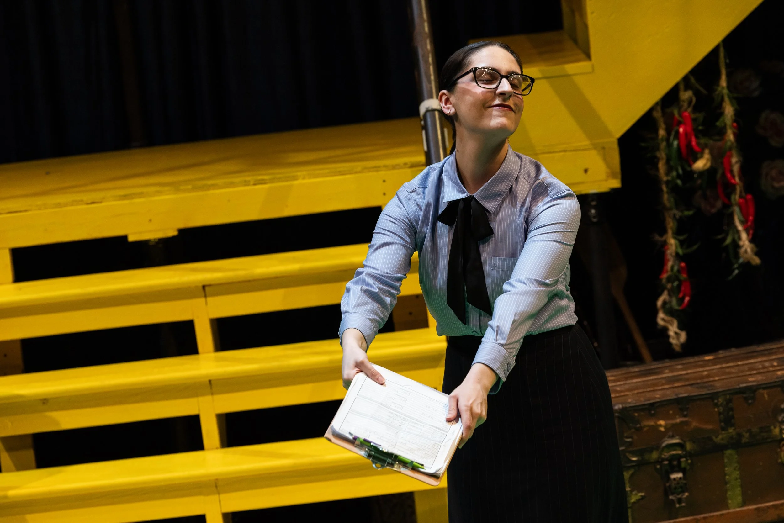 A woman with glasses, wearing a light blue pinstripe shirt with a black necktie, holding a clipboard, standing in front of yellow bleachers and decorated with festive greenery and red ribbons.