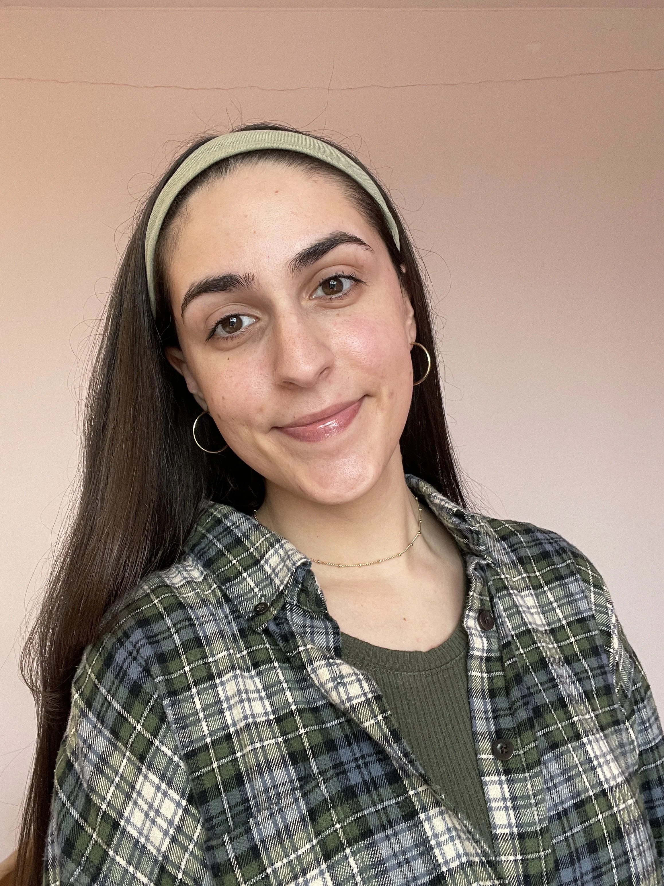 A young woman with brown hair, wearing a green headband, earrings, a plaid shirt over a green top, and a gold necklace, smiling at the camera against a plain beige wall.