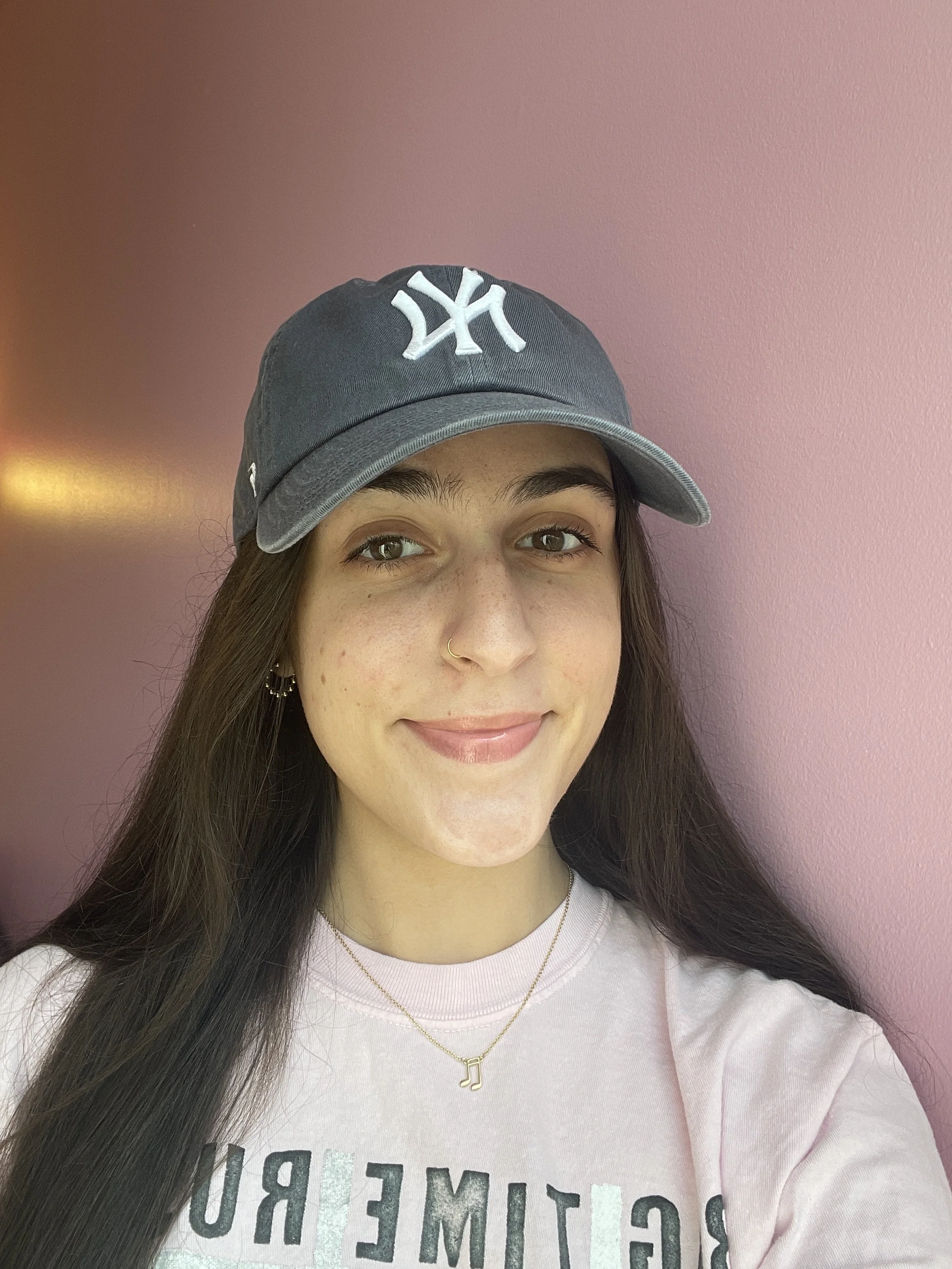A young woman with long dark hair, wearing a gray baseball cap with a white logo, gold hoop earrings, and a gold necklace with a musical note pendant, smiling against a pink wall background.